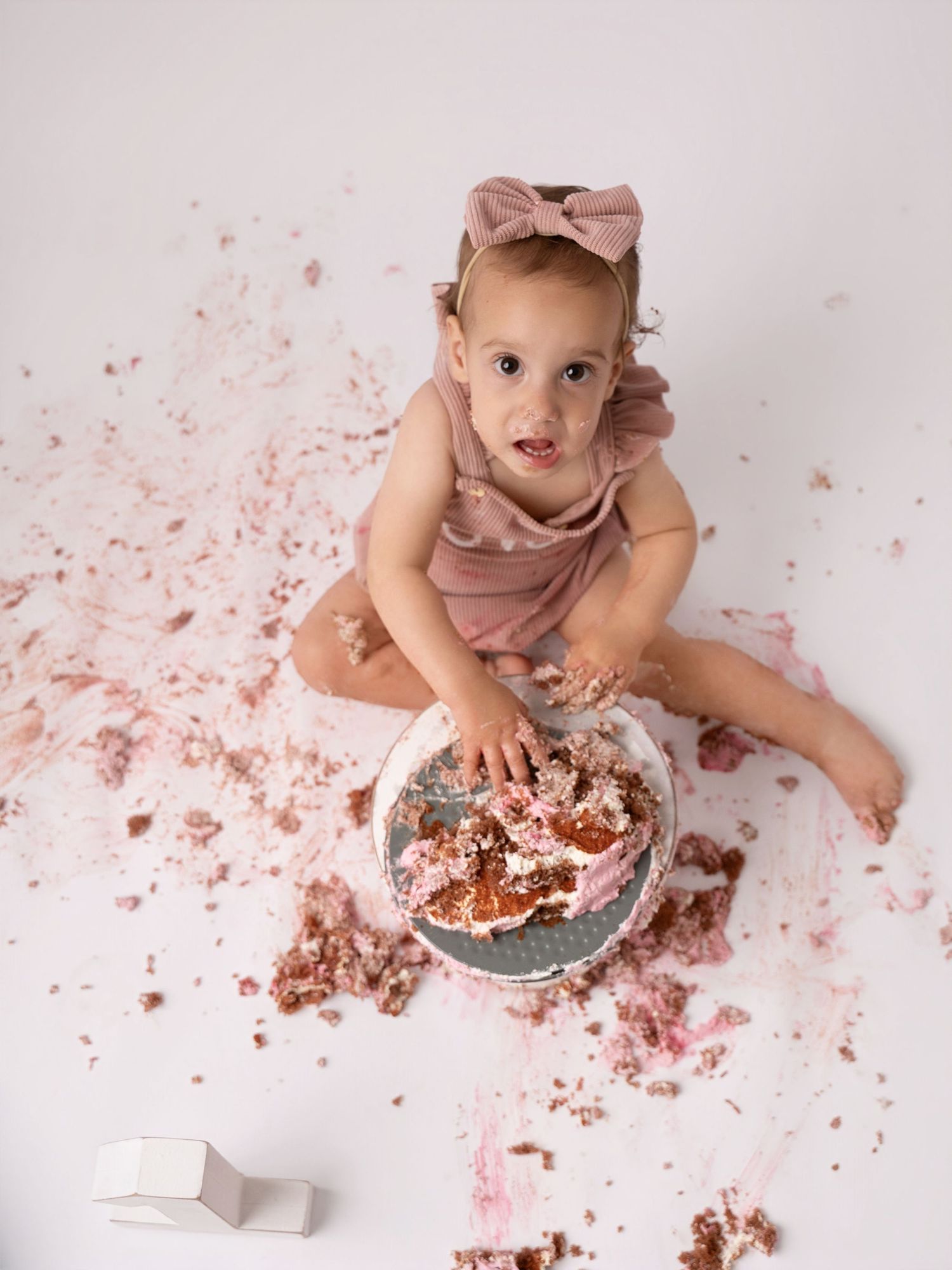 Baby girl in pink romper smashing her first birthday cake on white backdrop during cake smash photo session.