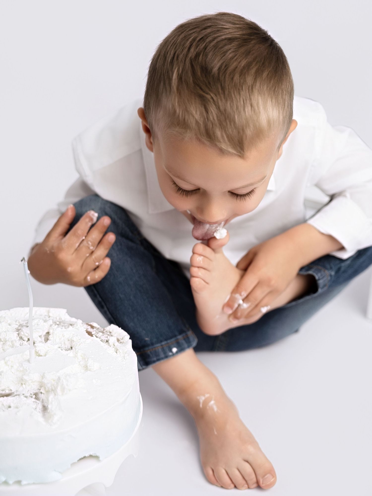 Young boy in white shirt and jeans licking frosting off his foot beside a white smash cake.