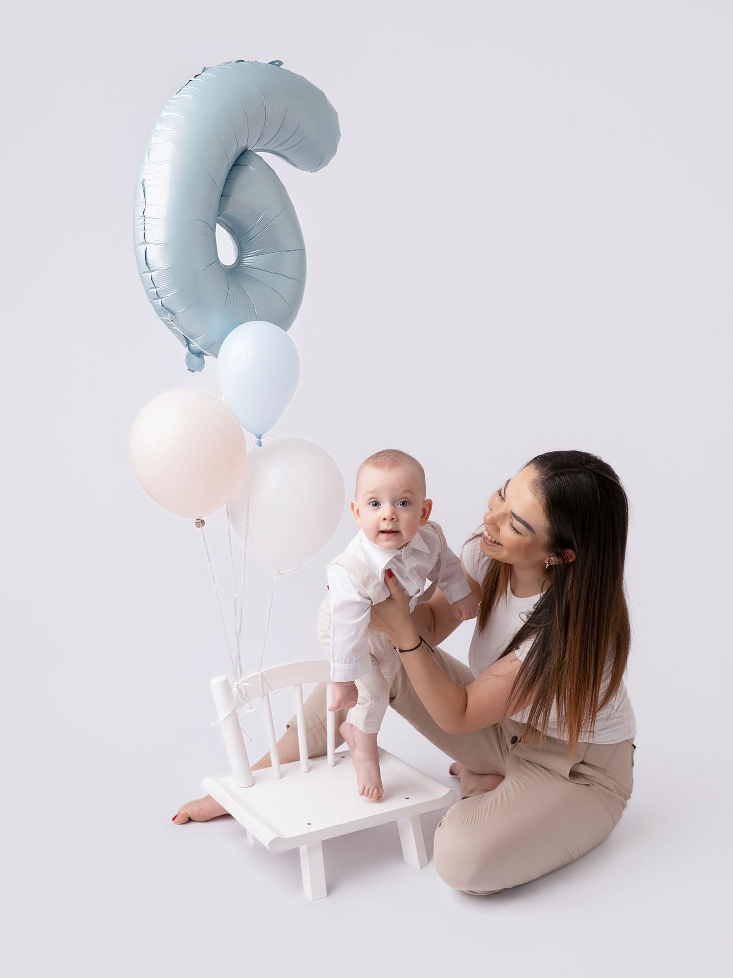 Mother holds smiling baby on white chair with blue number 6 balloon and pastel balloons for 6-month birthday photo.