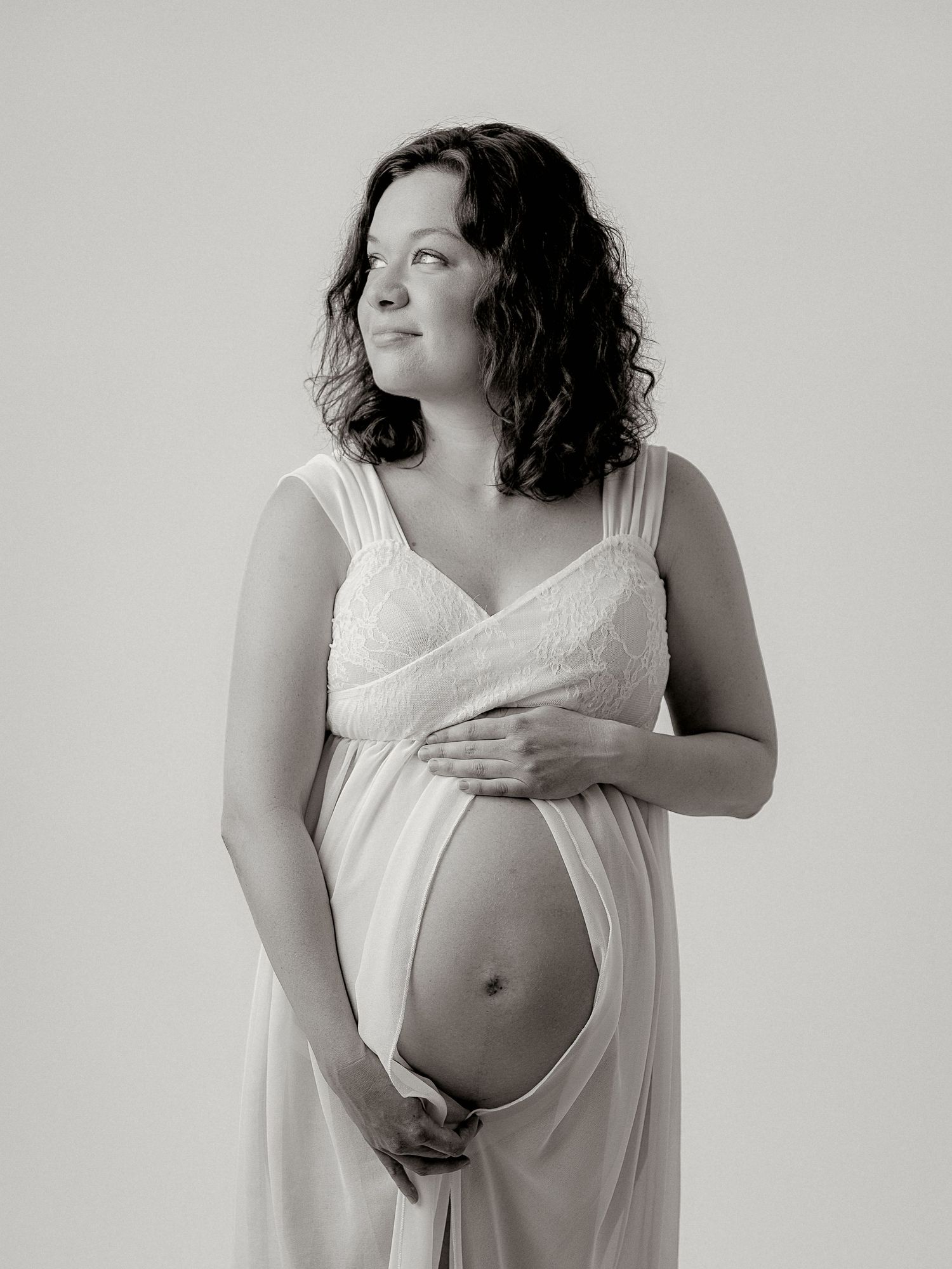 Black and white maternity photo of pregnant woman in white dress cradling her baby bump in a studio.