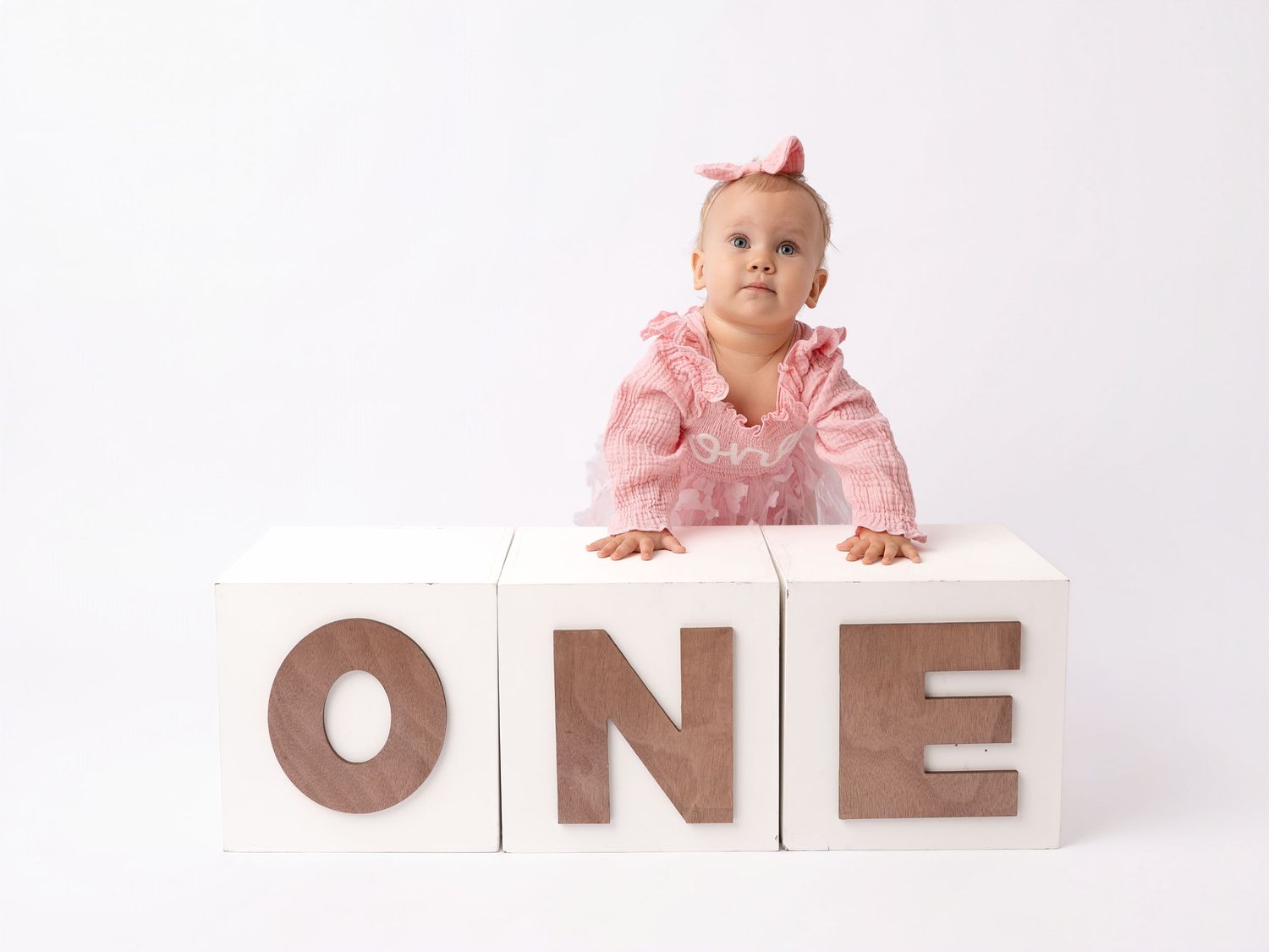 Baby girl in pink dress poses behind white blocks spelling ONE for her first birthday photo.