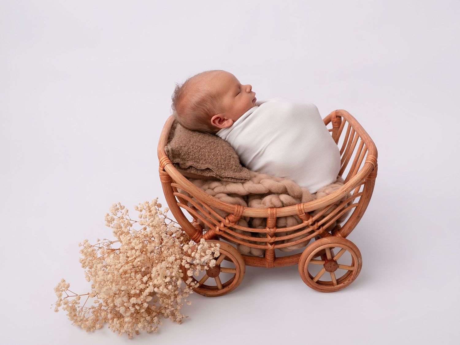 Newborn baby swaddled in white wrap sleeping in a small rattan pram with dried flowers on white background.