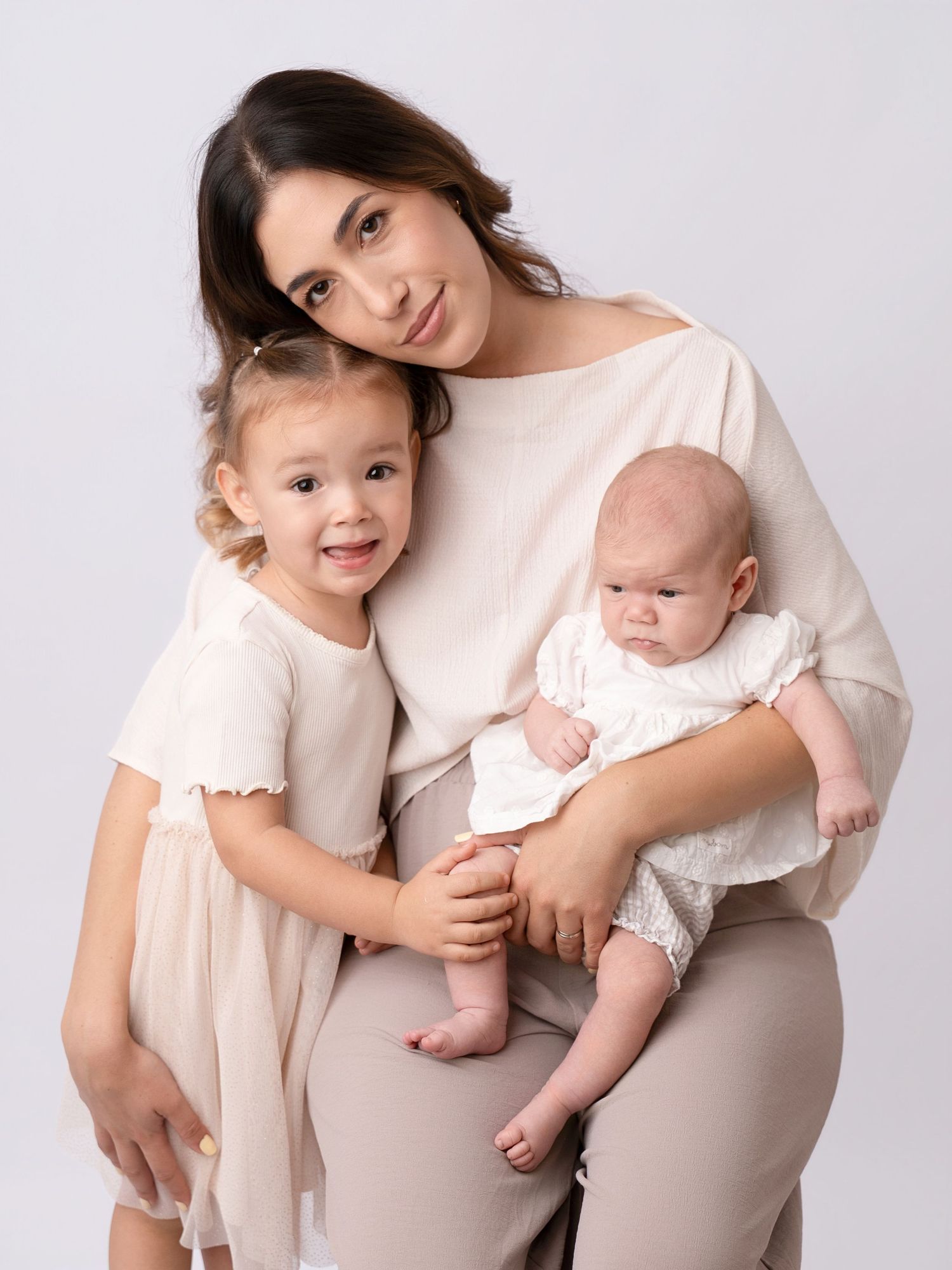 A mother poses with her toddler daughter and newborn baby in a soft, neutral-toned studio portrait.