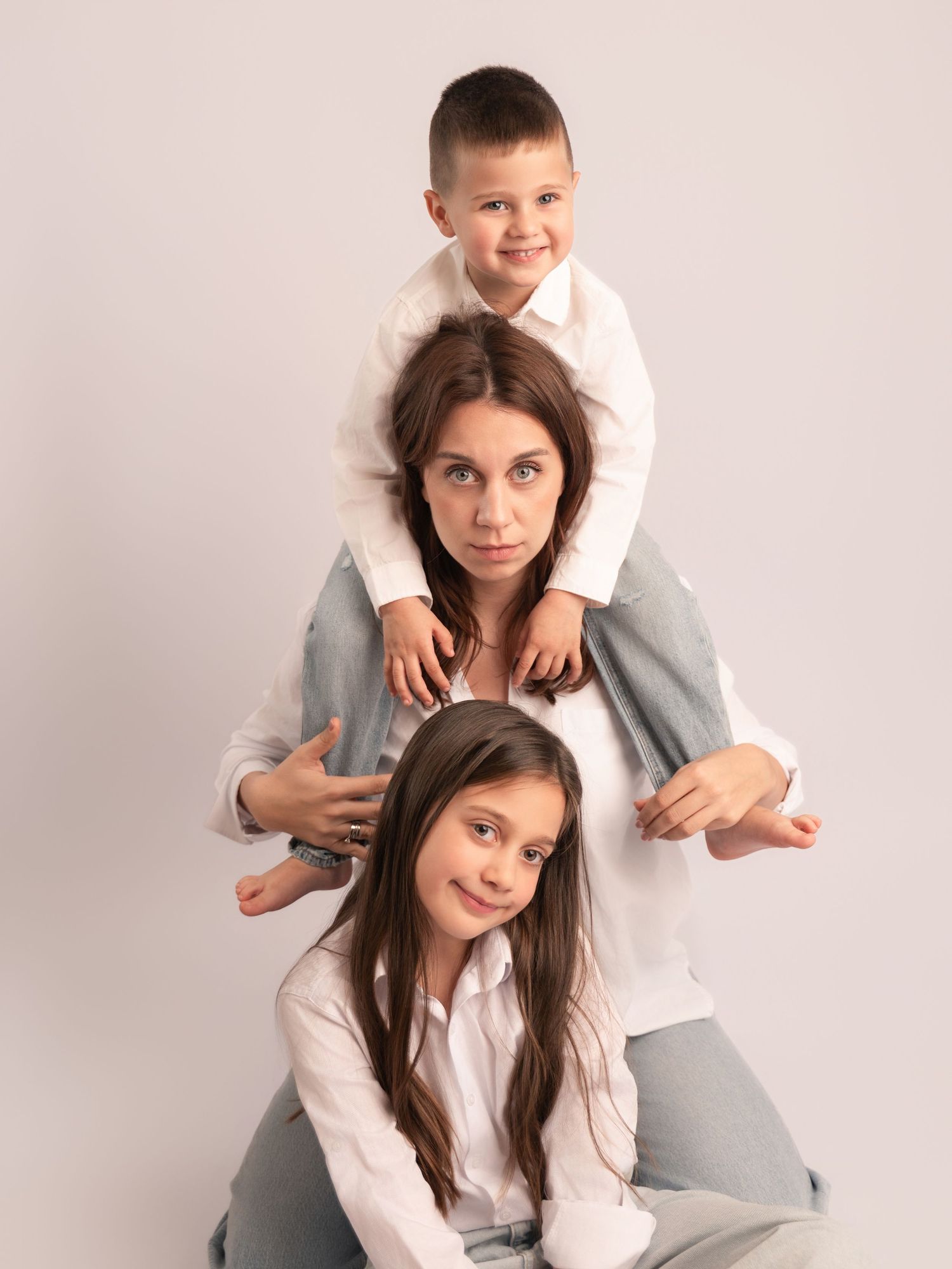 Mother with two children stacked in playful pyramid pose, all wearing white shirts in studio.