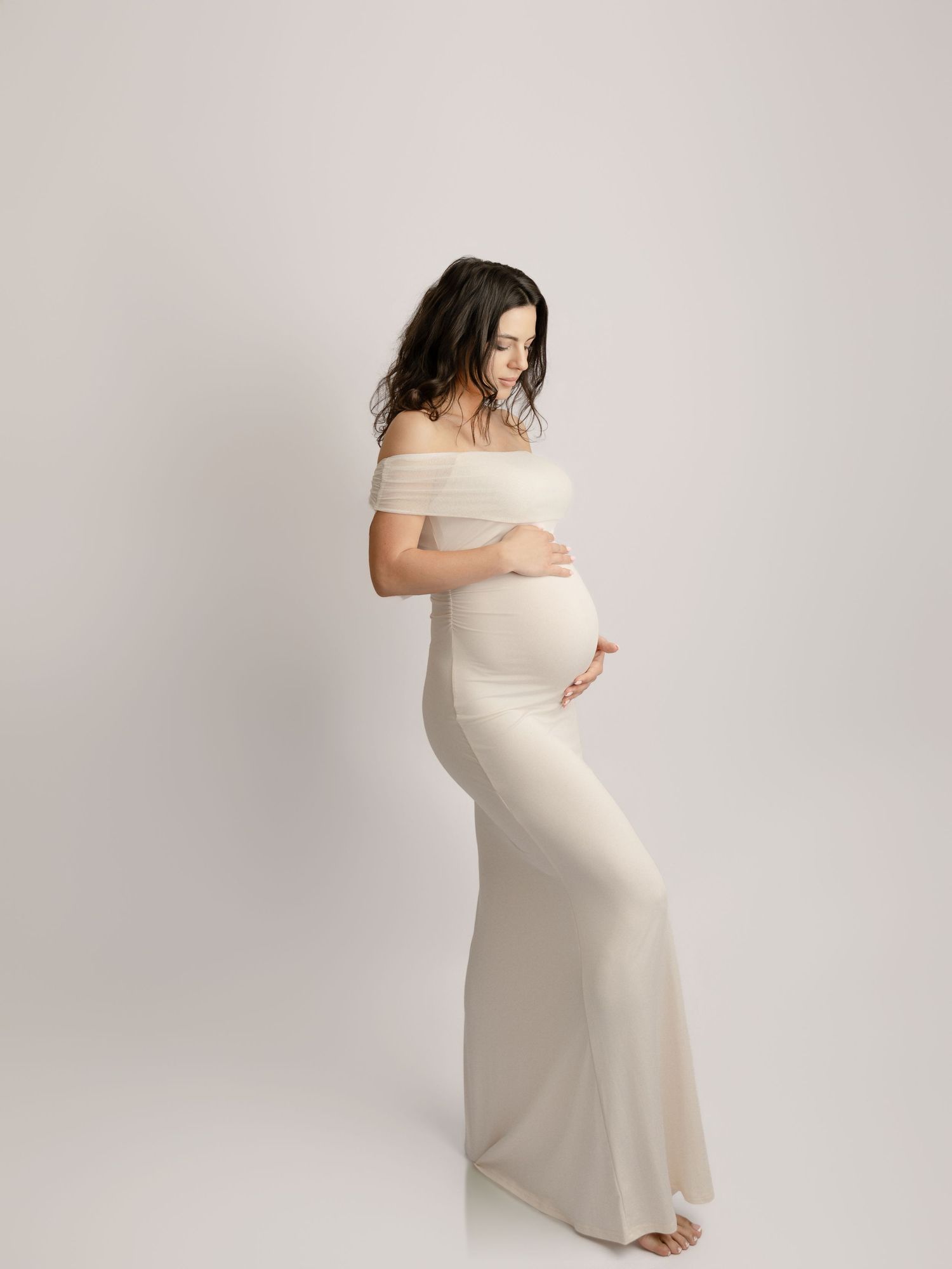 Pregnant woman in elegant off-shoulder cream maternity gown standing sideways.