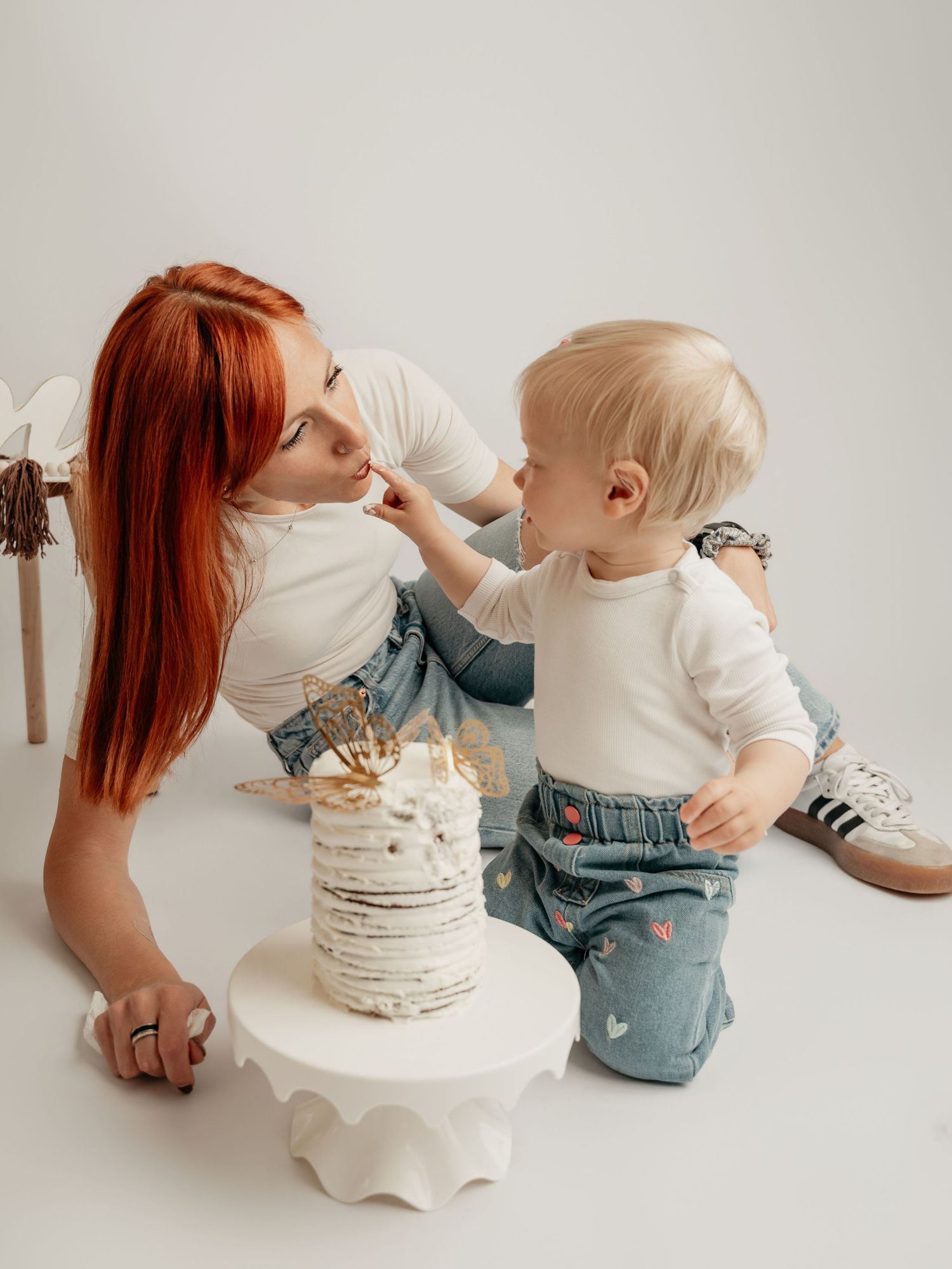 A red-haired mother smiles as her blonde toddler offers her a taste of birthday cake.