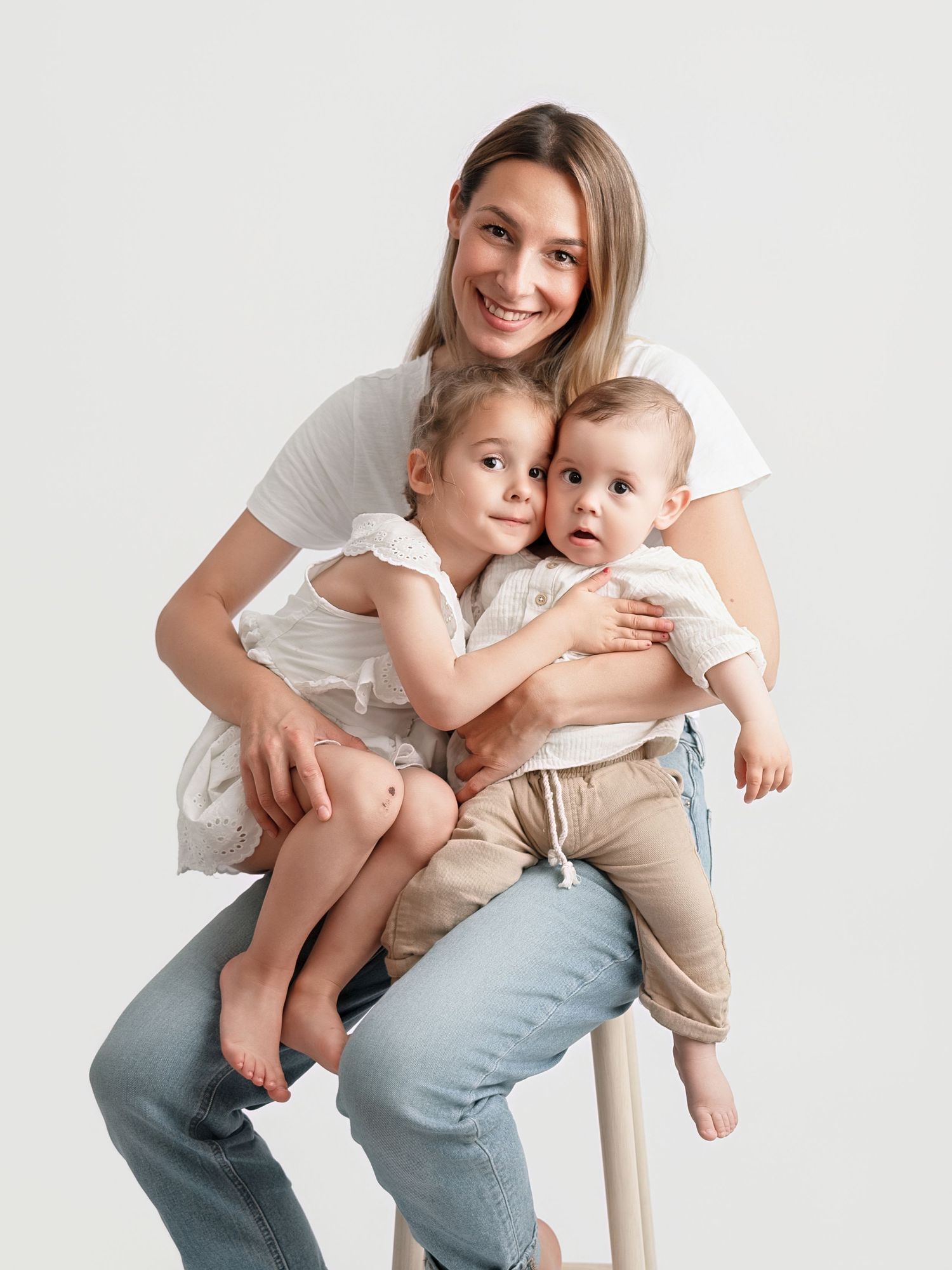 A smiling mother sits on a stool hugging her young daughter and baby in a bright studio portrait.