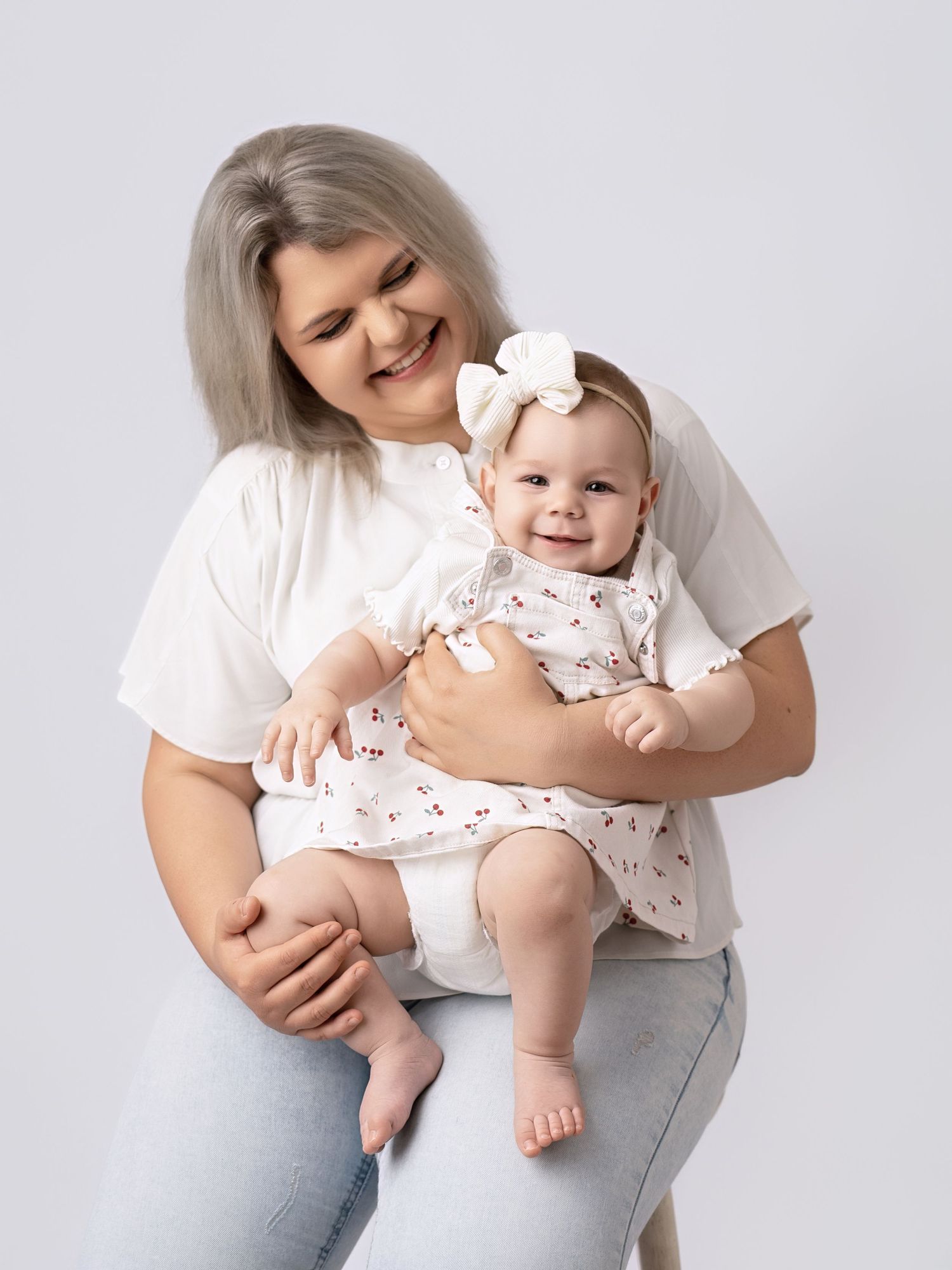 Smiling mother in white t-shirt holding happy baby girl in floral dress with white bow headband.