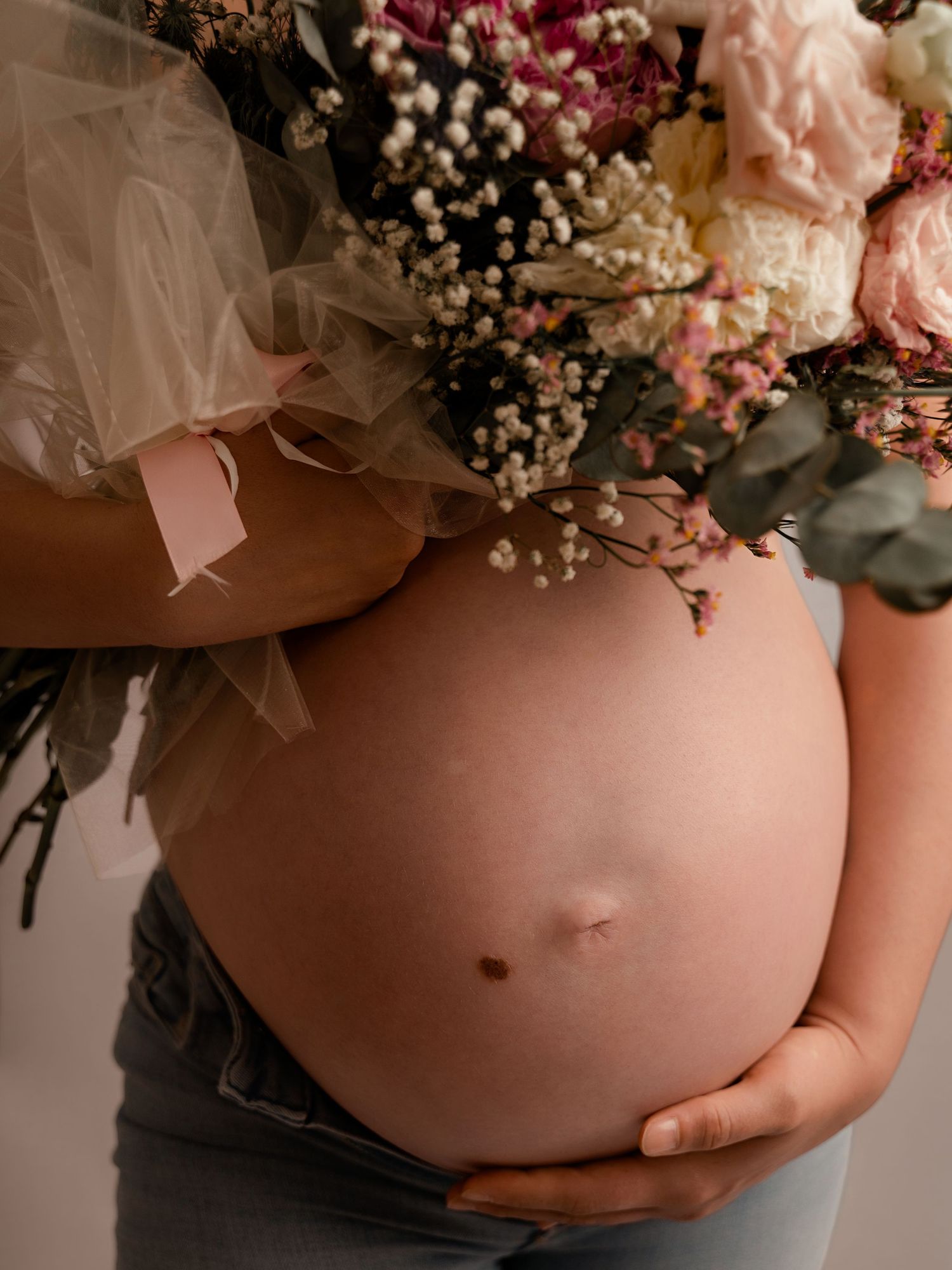 Pregnant woman cradling baby bump while holding a bouquet of pink and white flowers.