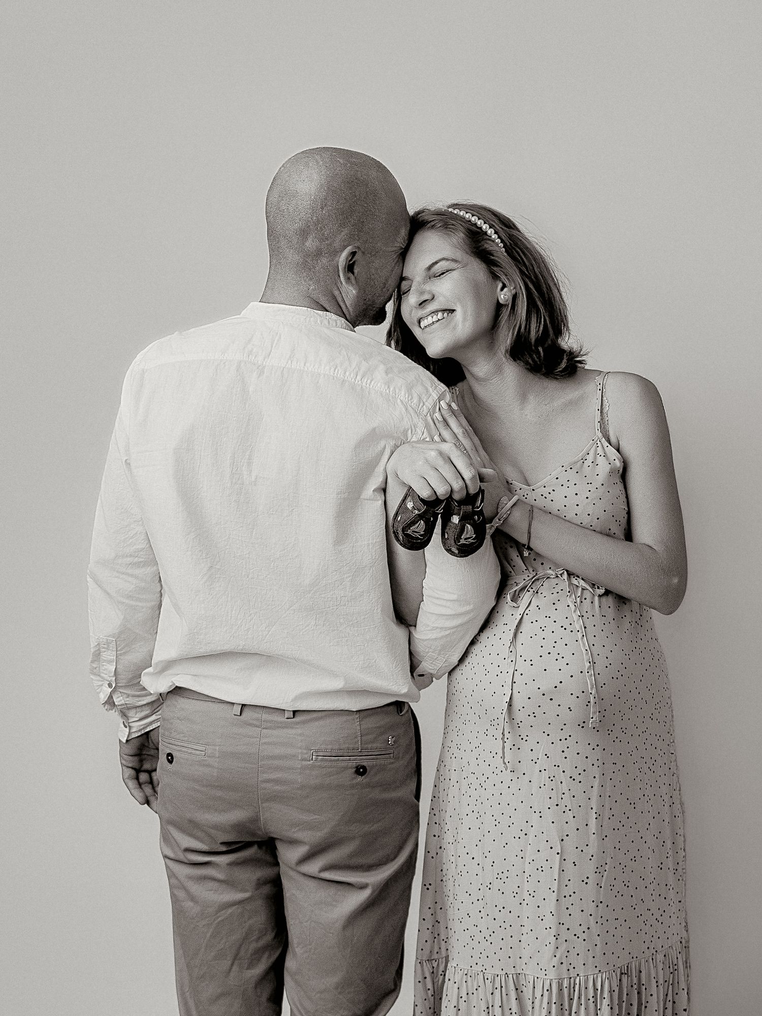 Black and white photo of smiling pregnant woman leaning on partners shoulder holding baby shoes.