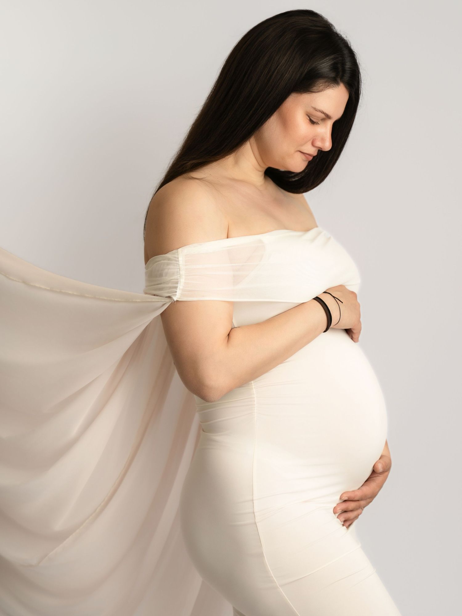 Pregnant woman in white off-shoulder dress holds baby bump with flowing fabric in studio.
