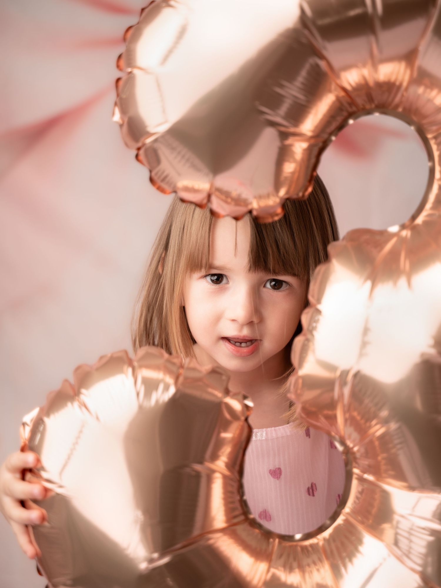 A little girl holds rose gold heart balloons close to her face in a warm-toned birthday photo session.