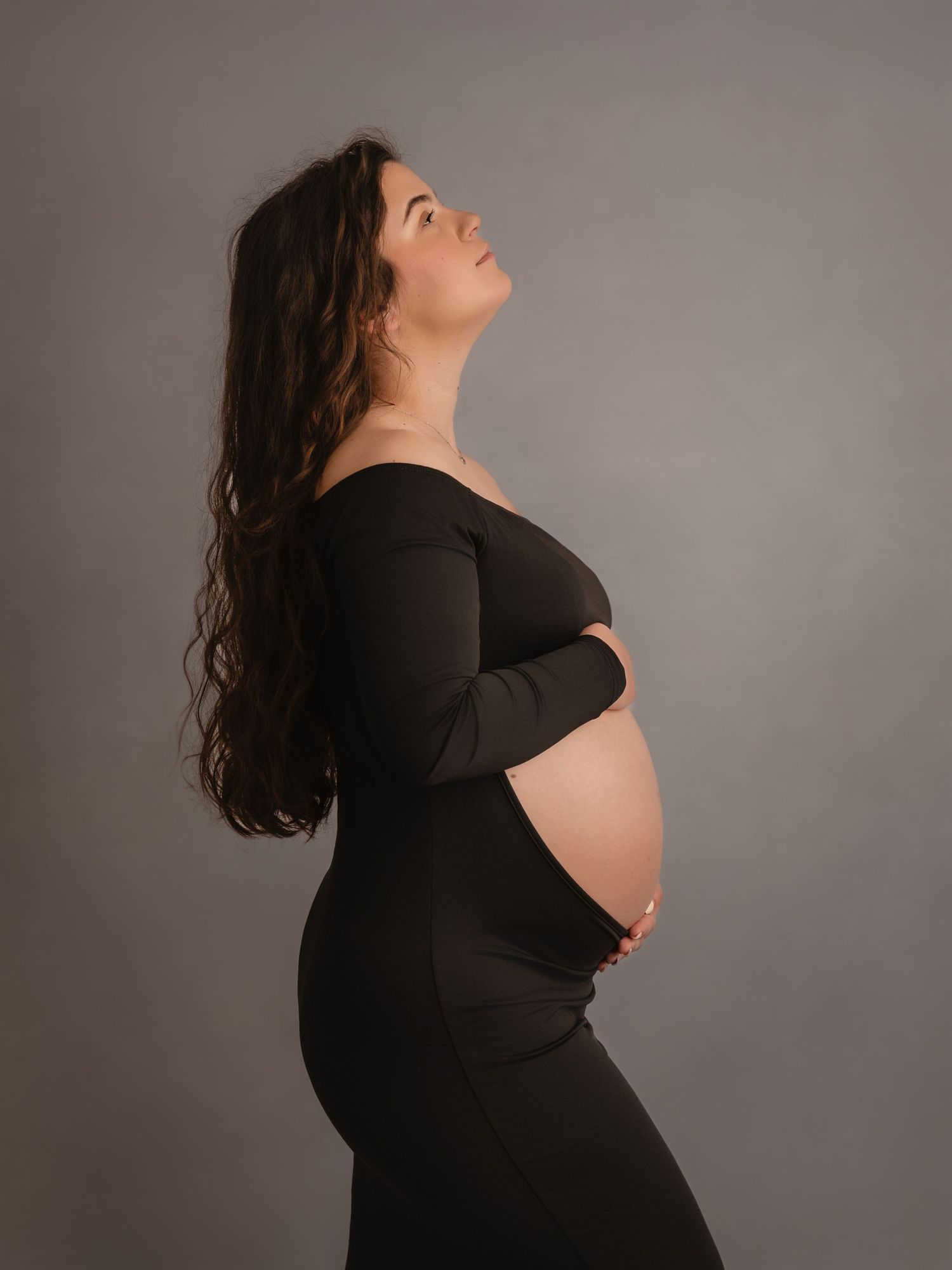 Pregnant woman in black off-shoulder maternity dress looking upward while cradling baby bump in studio.