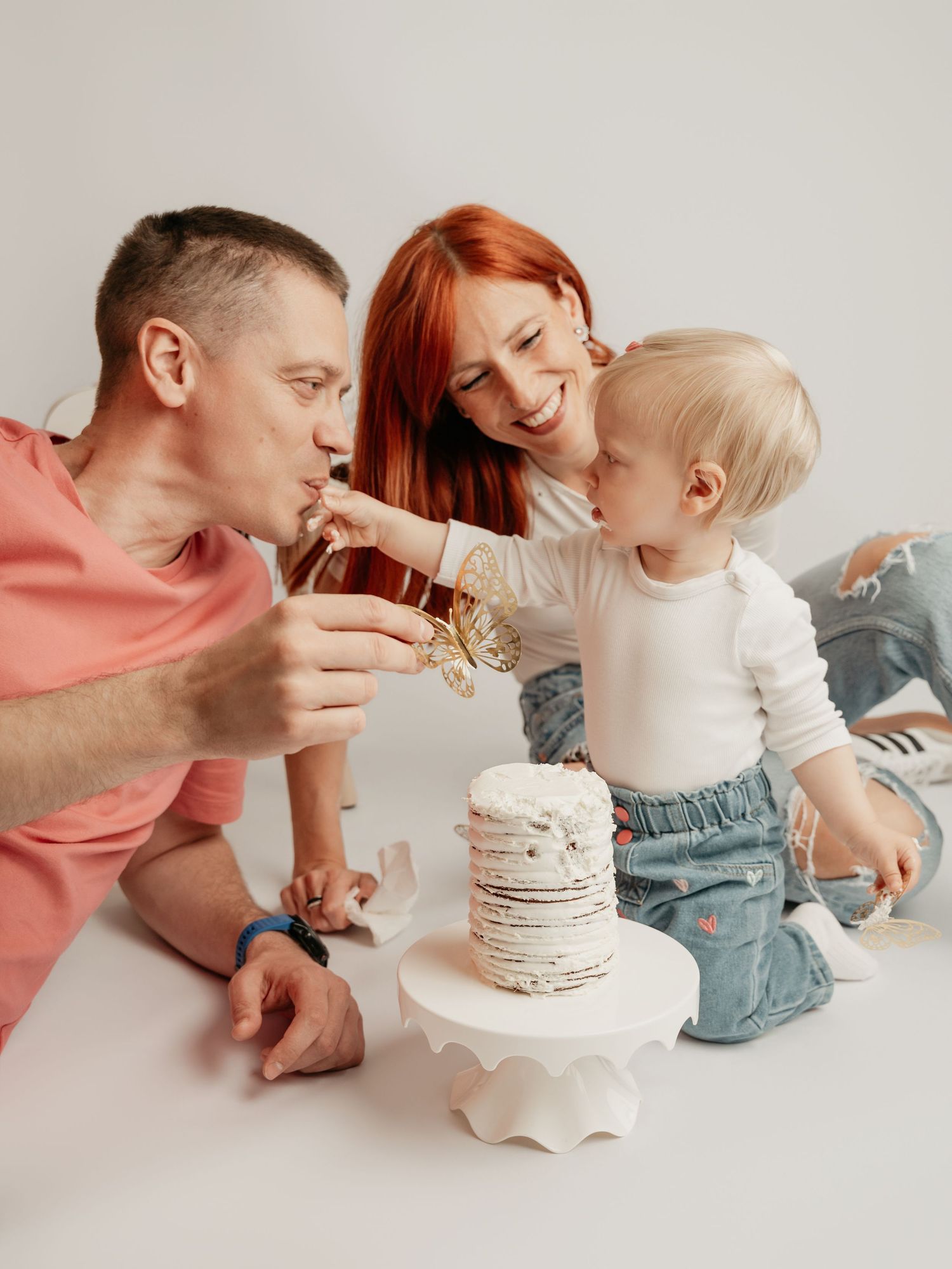 Parents laugh as their toddler shares birthday cake during a fun cake smash photo session.