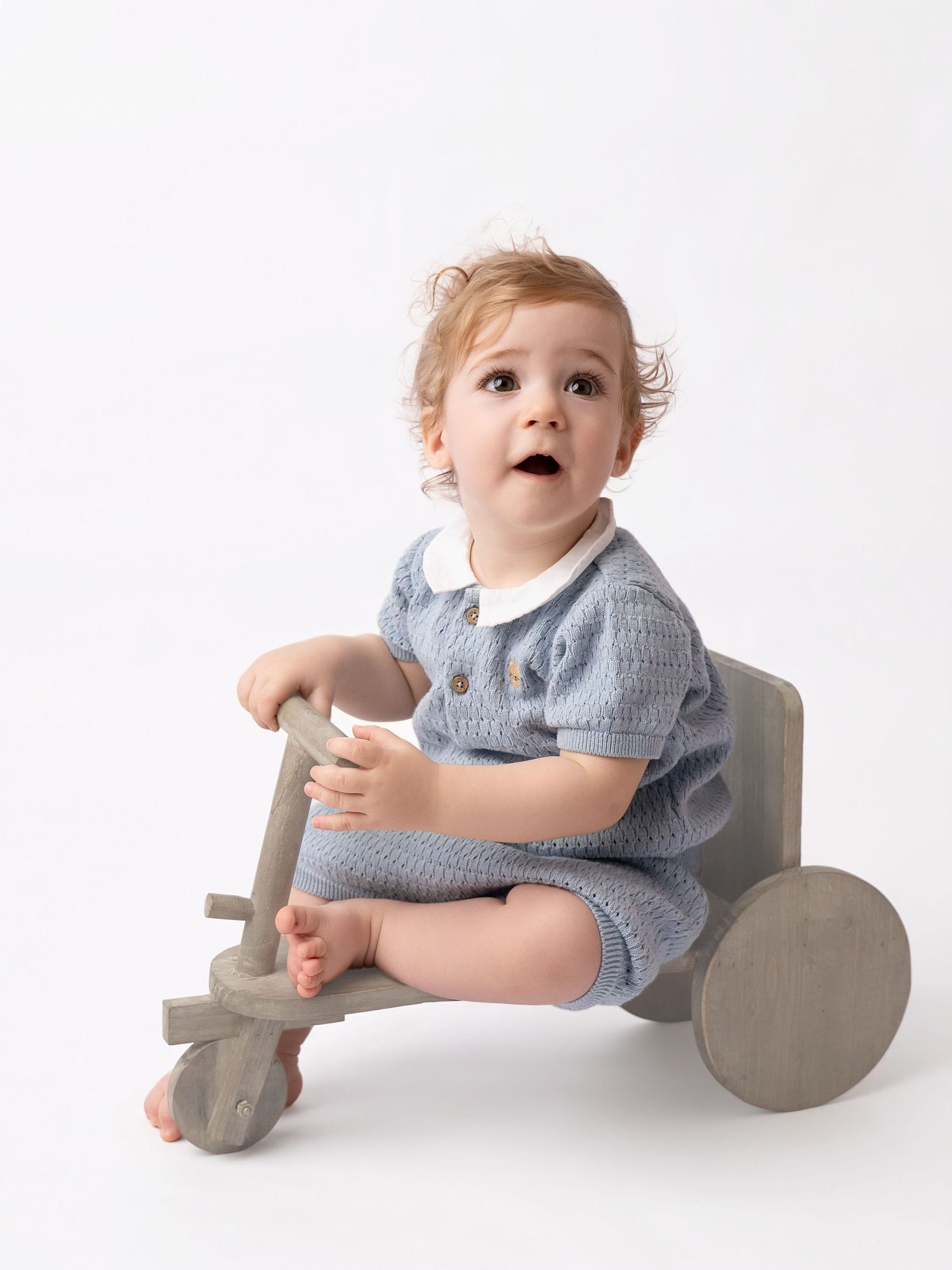 An adorable baby in a blue knit romper sits on a wooden toy tricycle against a clean white background.
