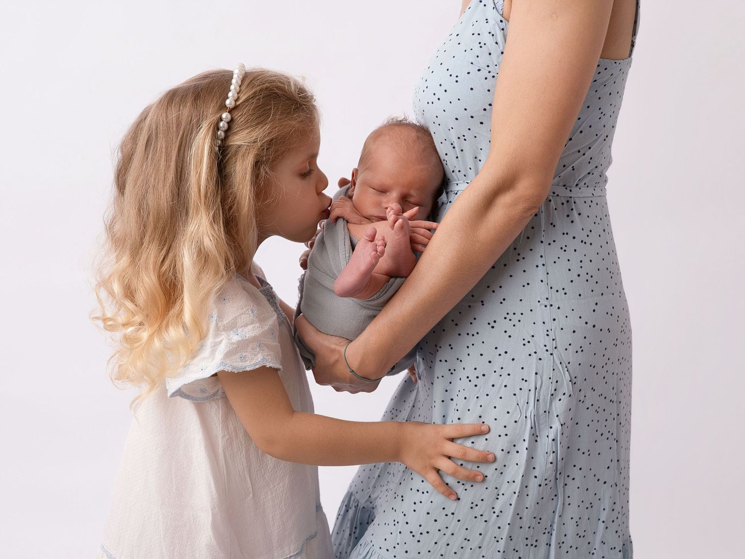 A young girl kisses a newborn baby held by a mother in a blue polka dot dress against a white background.