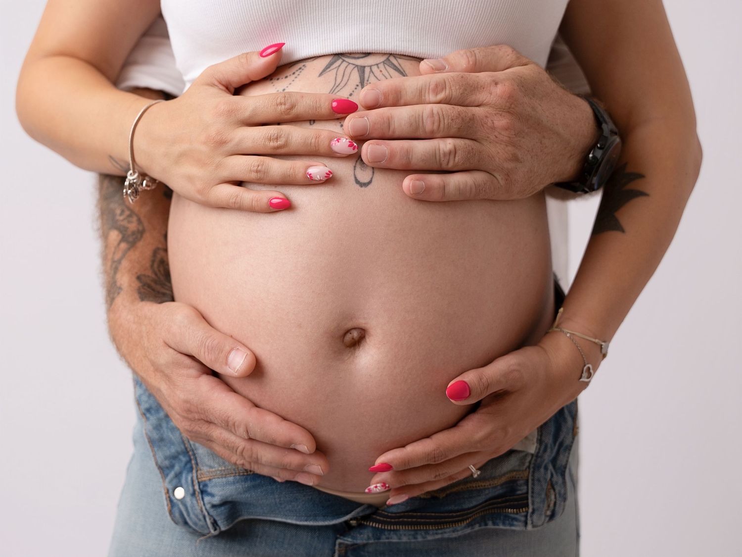 Close-up of pregnant belly with tattooed partner placing hands over expecting mother's bump.