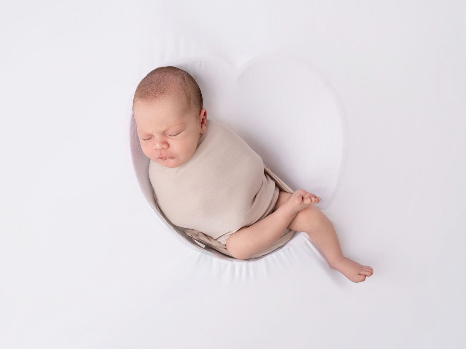 Newborn baby swaddled in beige wrap sleeping peacefully in a white bowl prop on a white background.