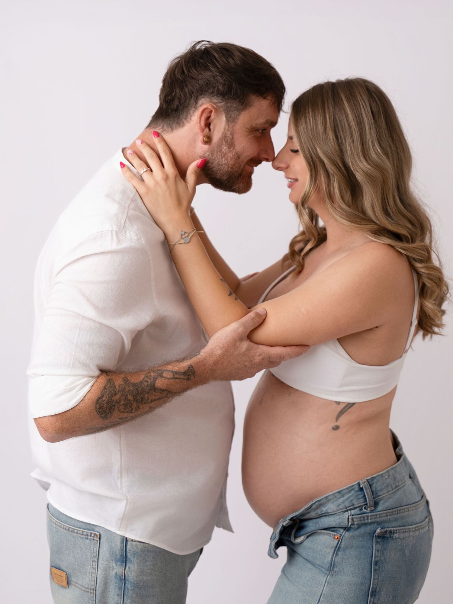 Couple sharing an intimate moment during a maternity photo shoot, pregnant woman in white crop top and jeans.