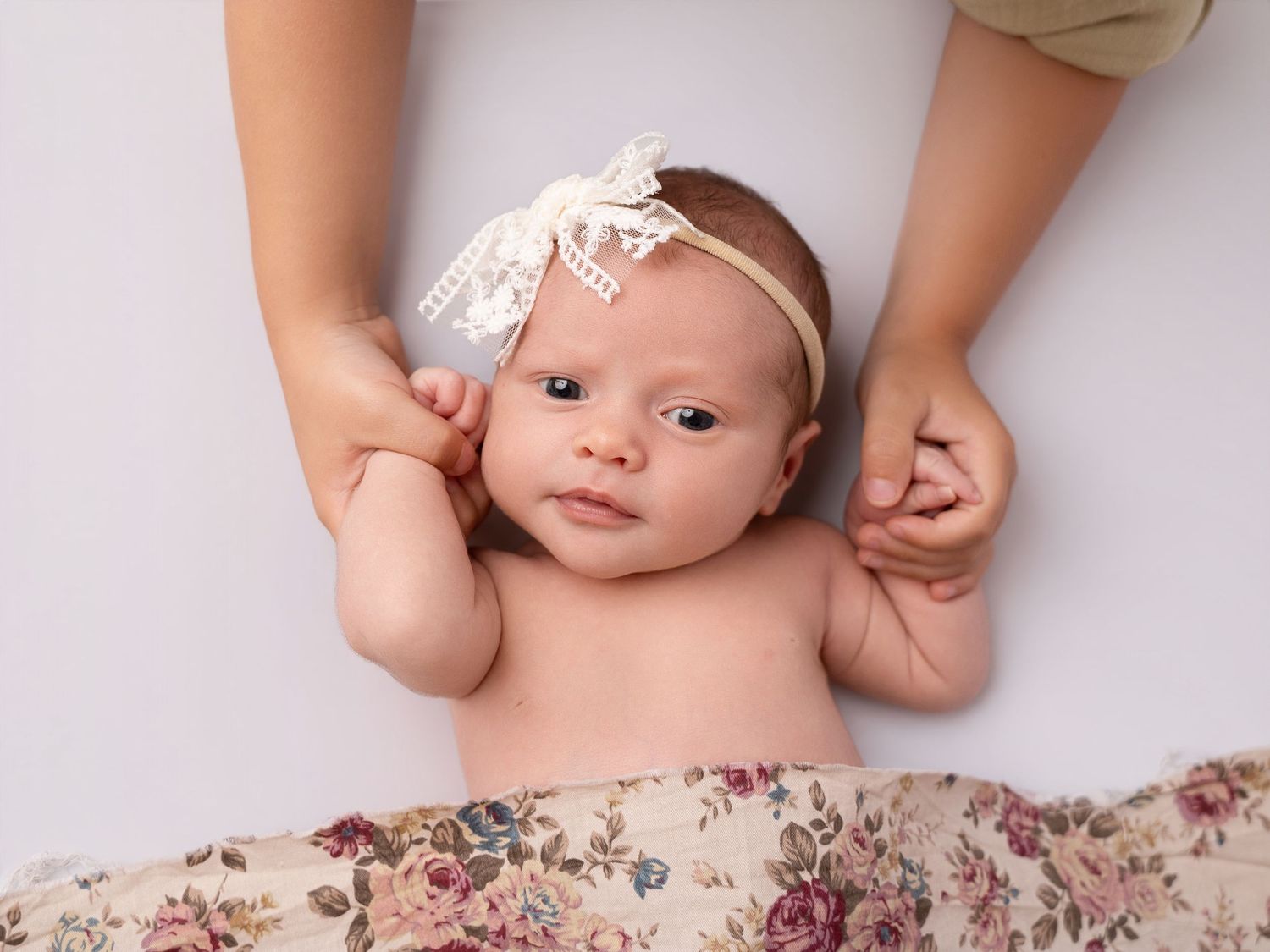 Newborn baby girl wearing a lace headband, lying on a floral wrap while an adult gently holds her hands.