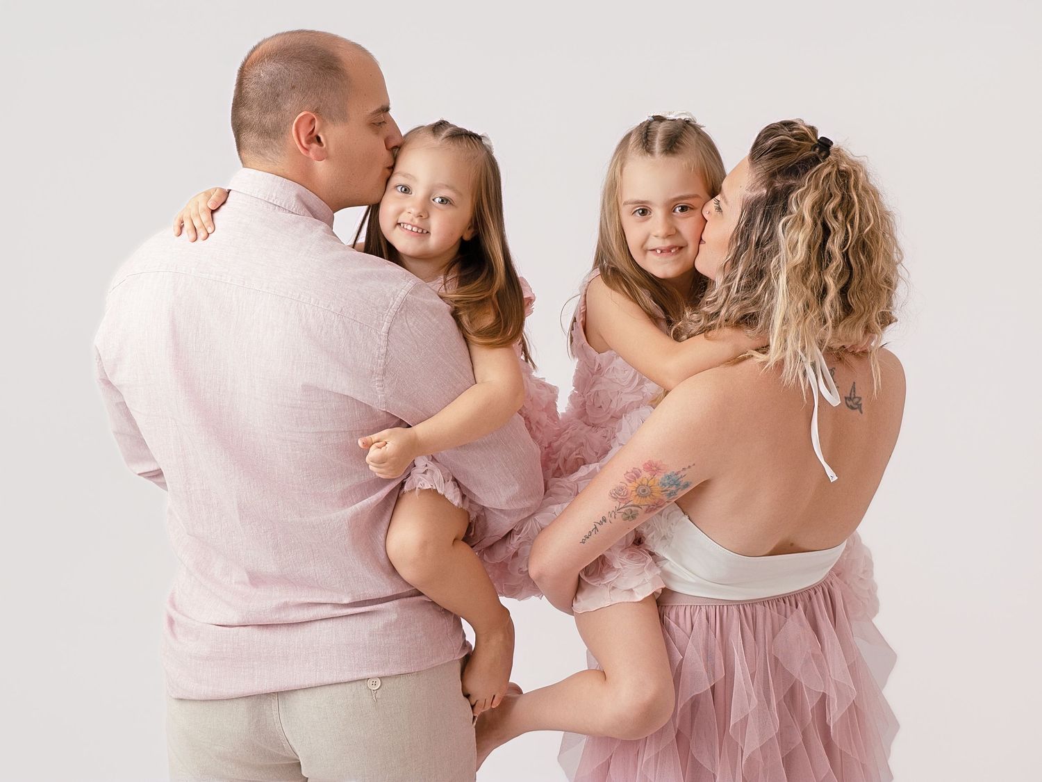Family portrait of parents holding two young girls in matching pink dresses against a white background.