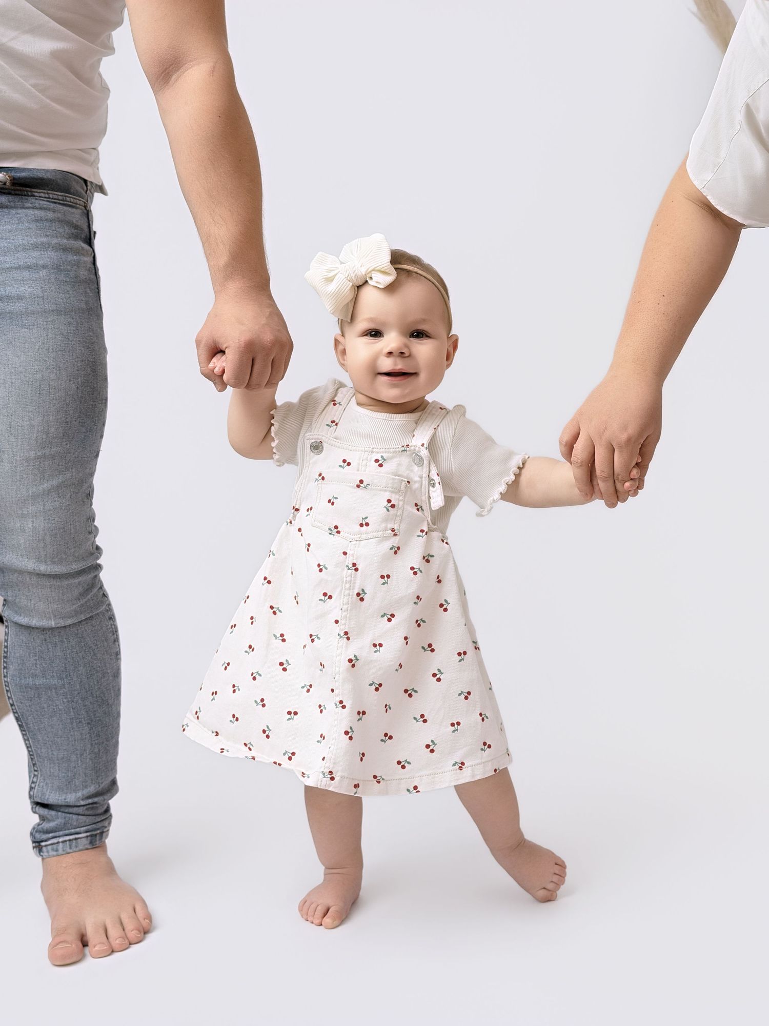 Smiling baby girl in cherry print dress and bow headband learning to walk with help from two adults.