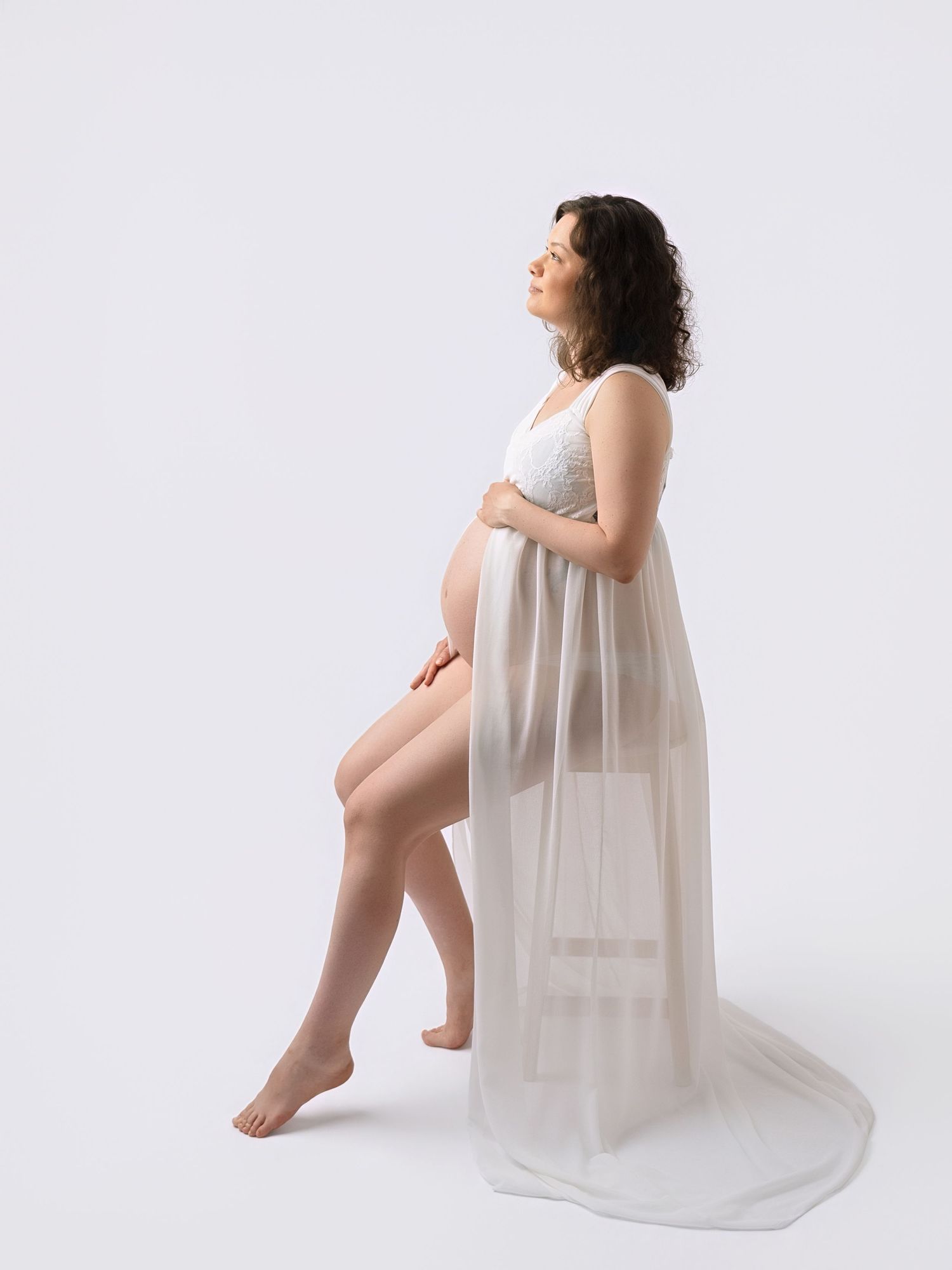 Pregnant woman in white lace bra and sheer flowing skirt poses gracefully against white background.
