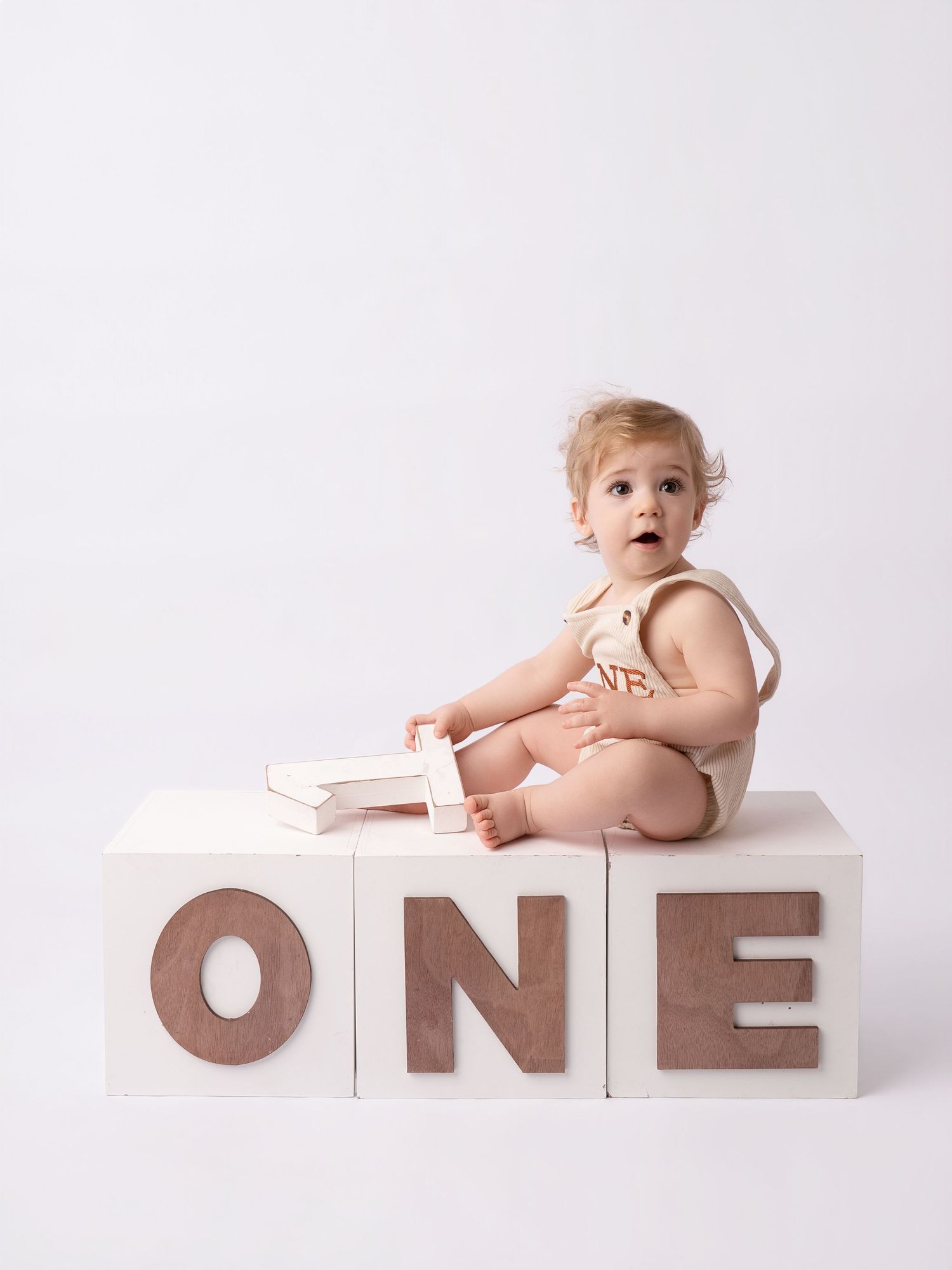 Cute baby sitting on white ONE letter blocks in beige romper for first birthday photos.