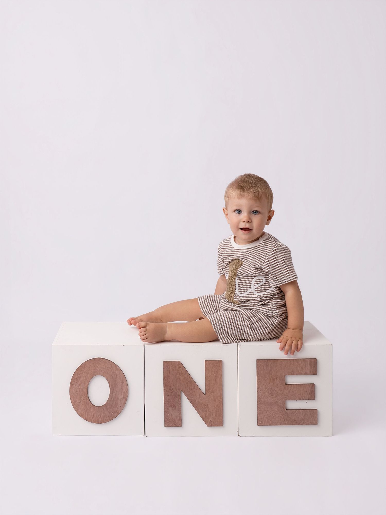 Smiling baby boy sitting on white ONE letter blocks, wearing a striped outfit, celebrating his first birthday.
