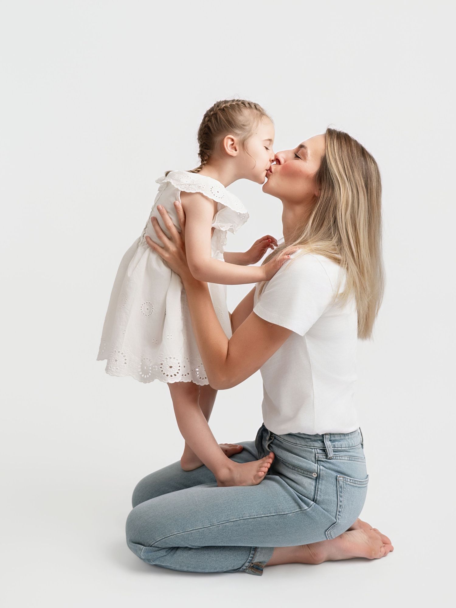 A mother kneeling on the floor kisses her toddler daughter in a white dress against a light background.
