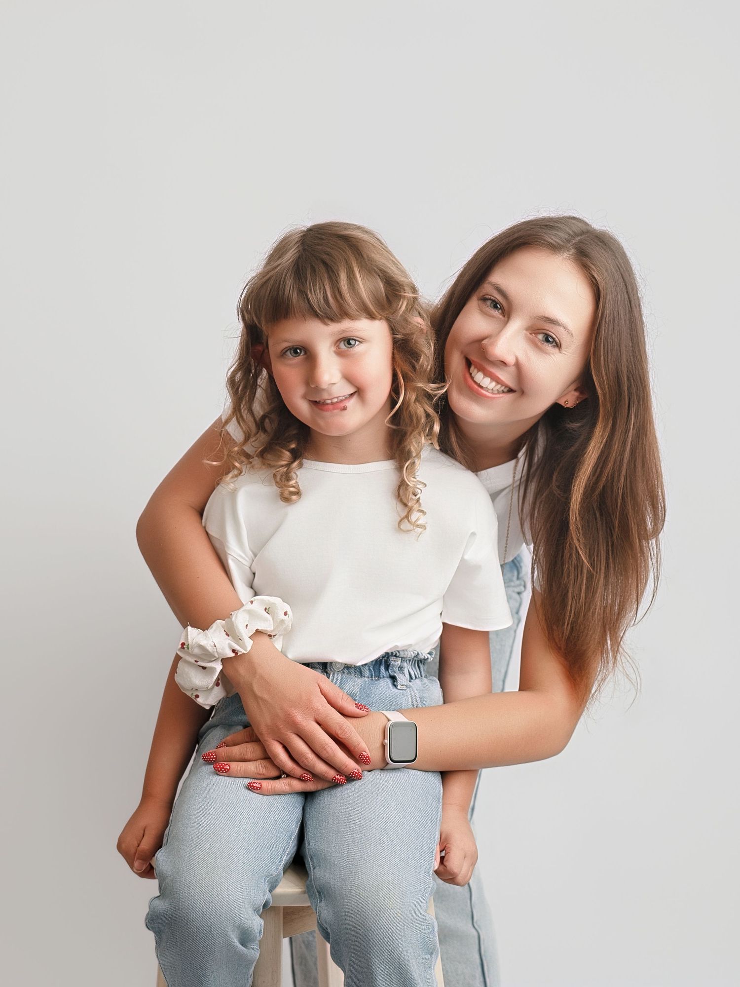 A young woman and little girl in matching white tees and jeans pose together smiling in a studio portrait.
