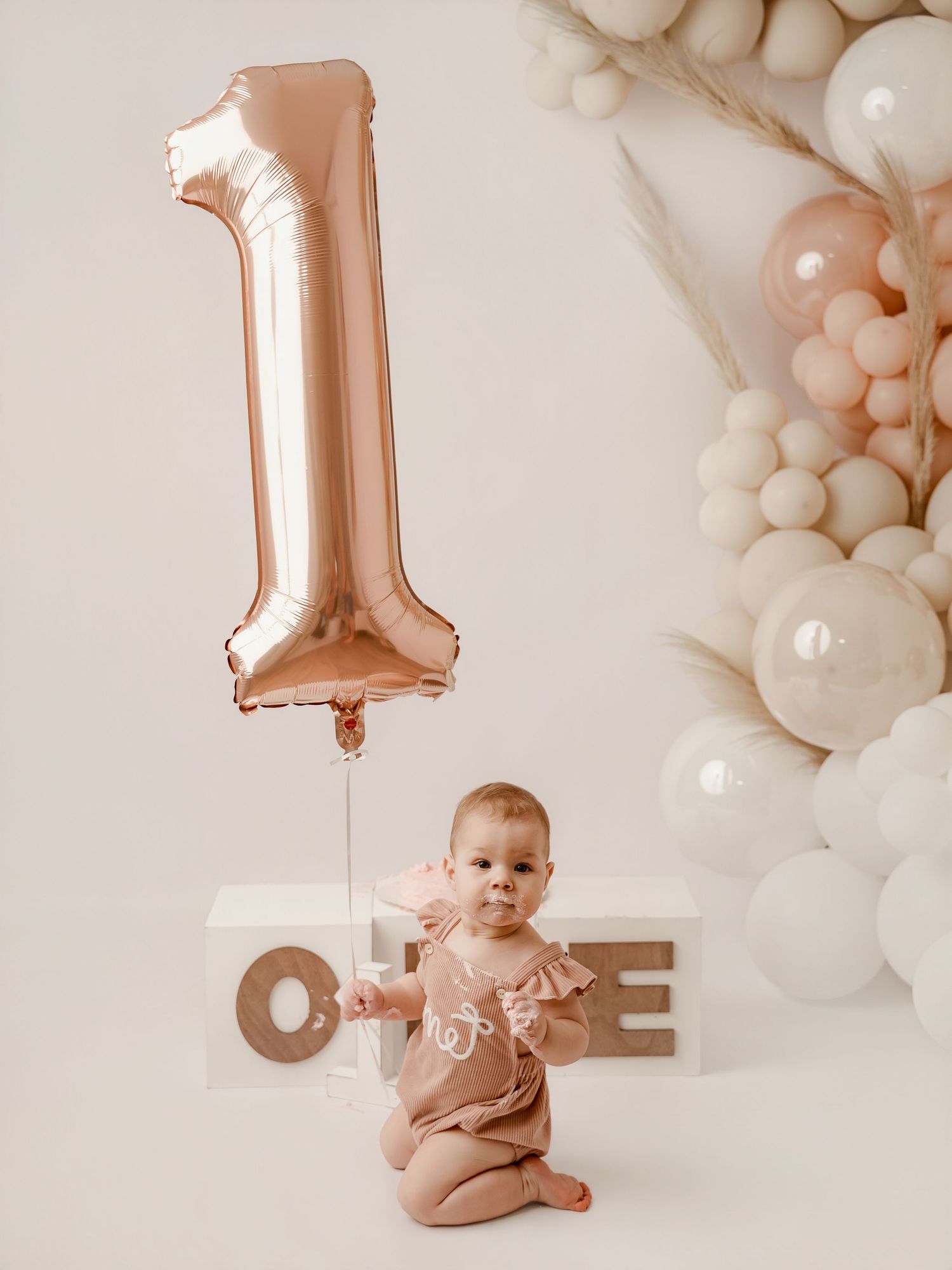Baby girl in pink romper sits before ONE blocks with rose gold number 1 balloon and pastel balloon arch.
