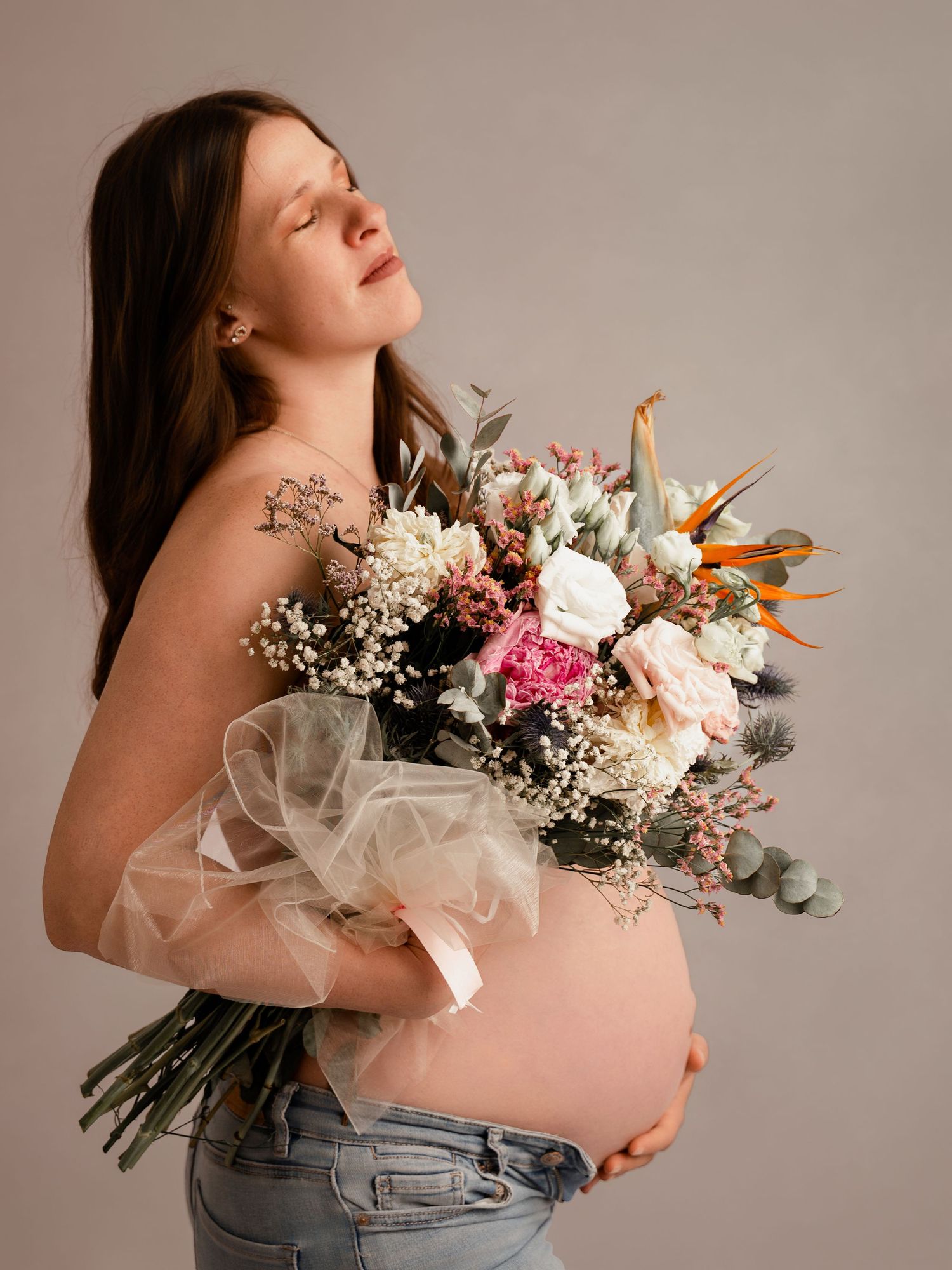 Pregnant woman holding a large floral bouquet with eyes closed, wearing jeans against a neutral background.