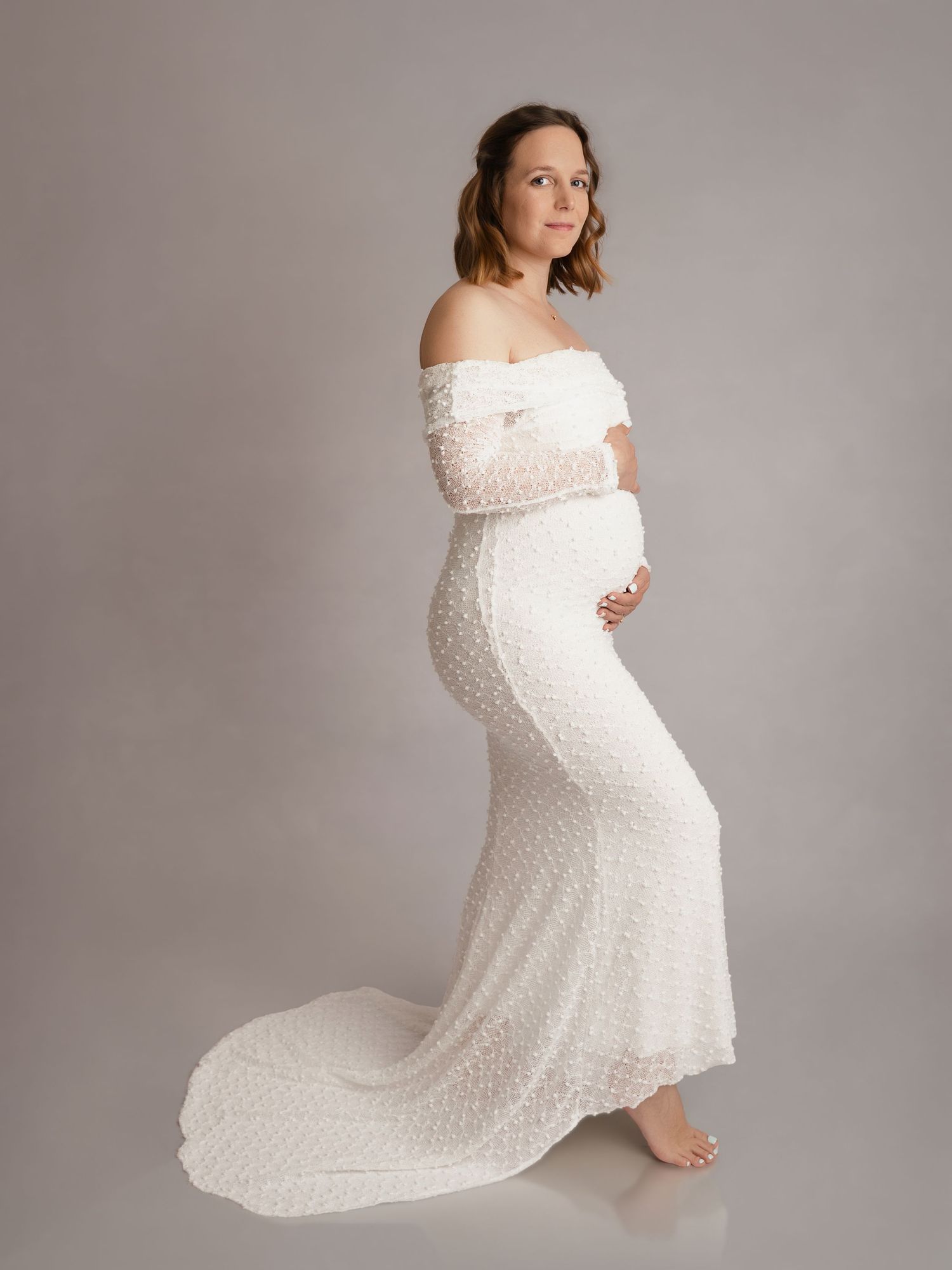 A pregnant woman in a stunning white lace off-shoulder gown poses gracefully in profile in a professional studio setting.