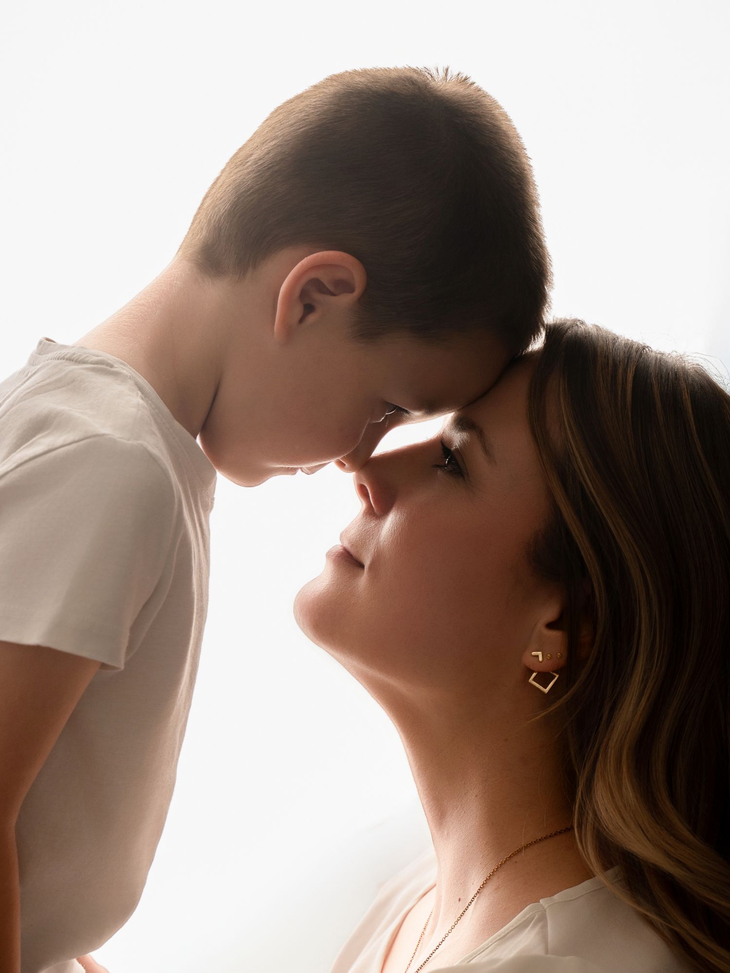 A mother and young son touch foreheads tenderly in a softly backlit studio portrait.
