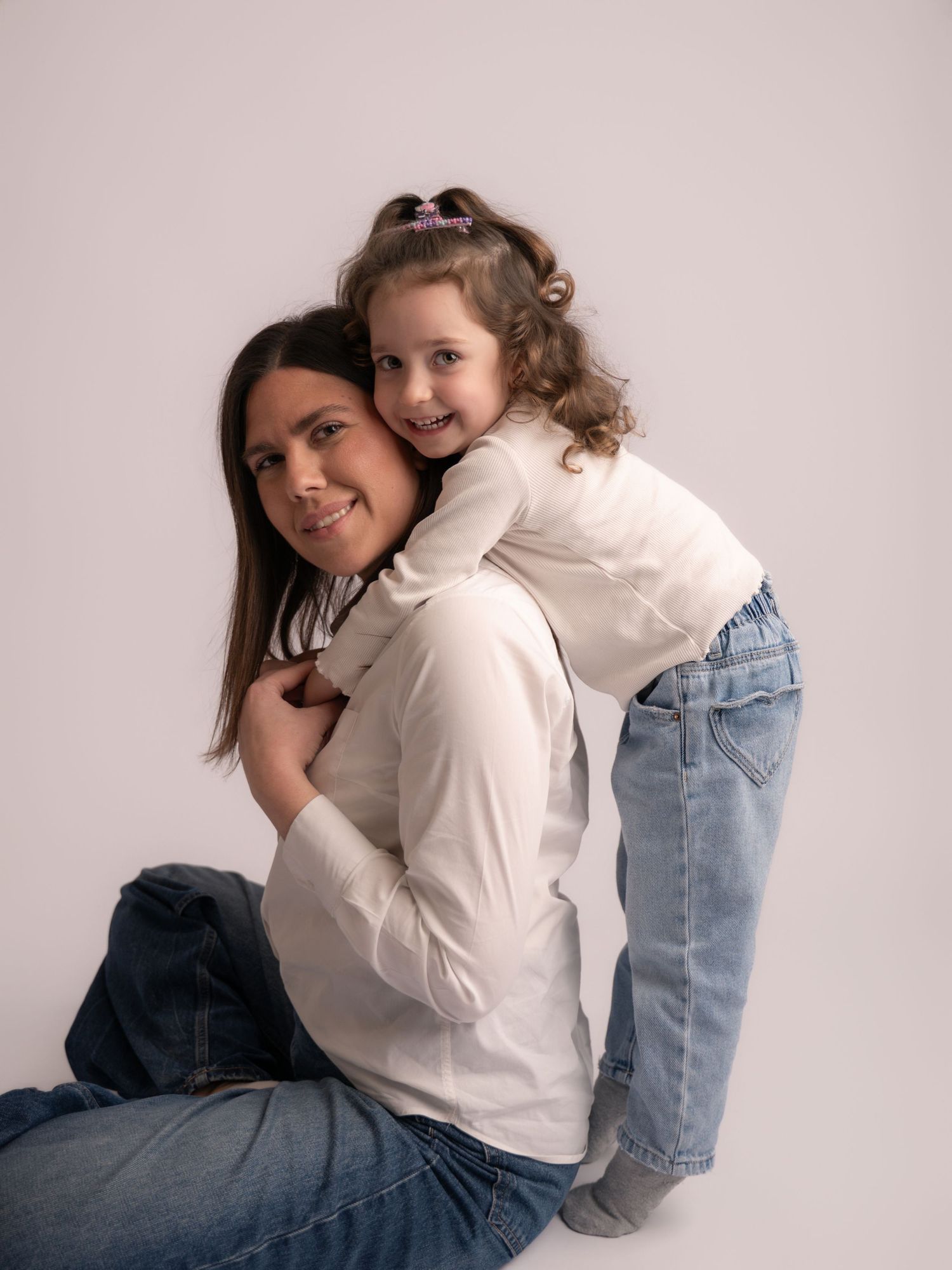 Mother and young daughter in matching white tops and jeans share a warm embrace in studio.