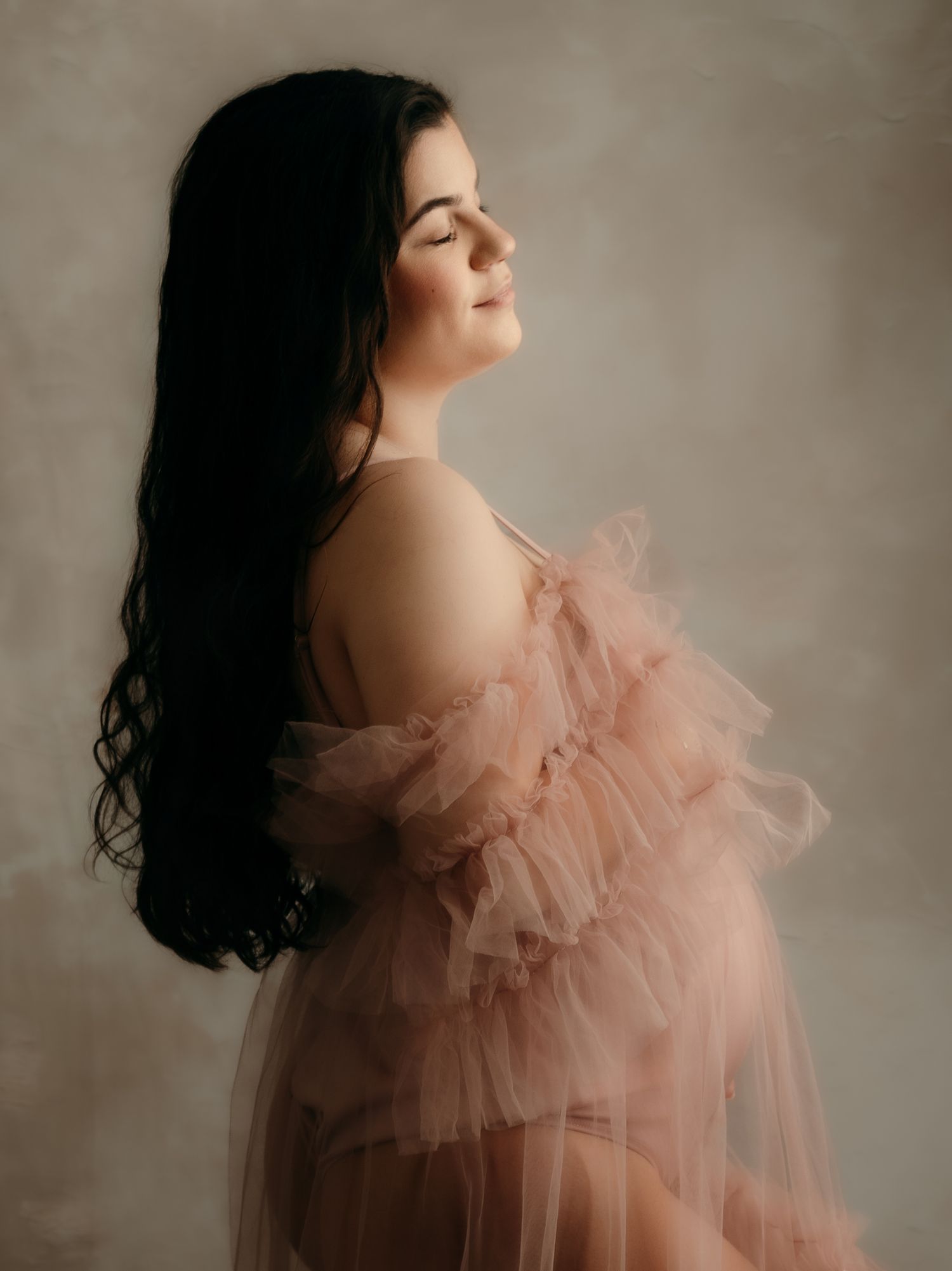 Side profile of a pregnant woman in a sheer pink ruffled tulle maternity dress with eyes closed peacefully.
