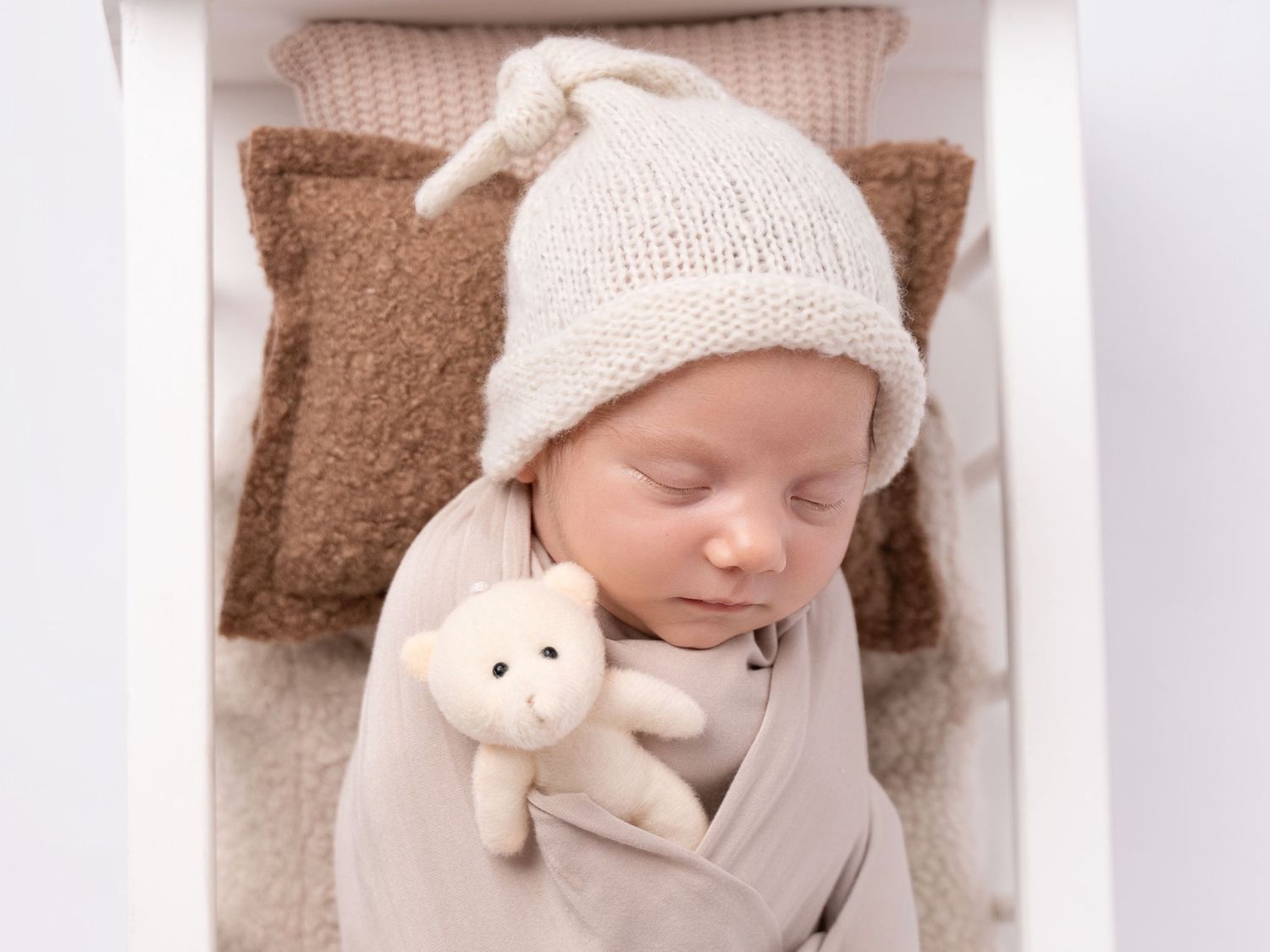 Sleeping newborn baby swaddled in gray wrap wearing a knit hat, holding a small white teddy bear in a white crib.