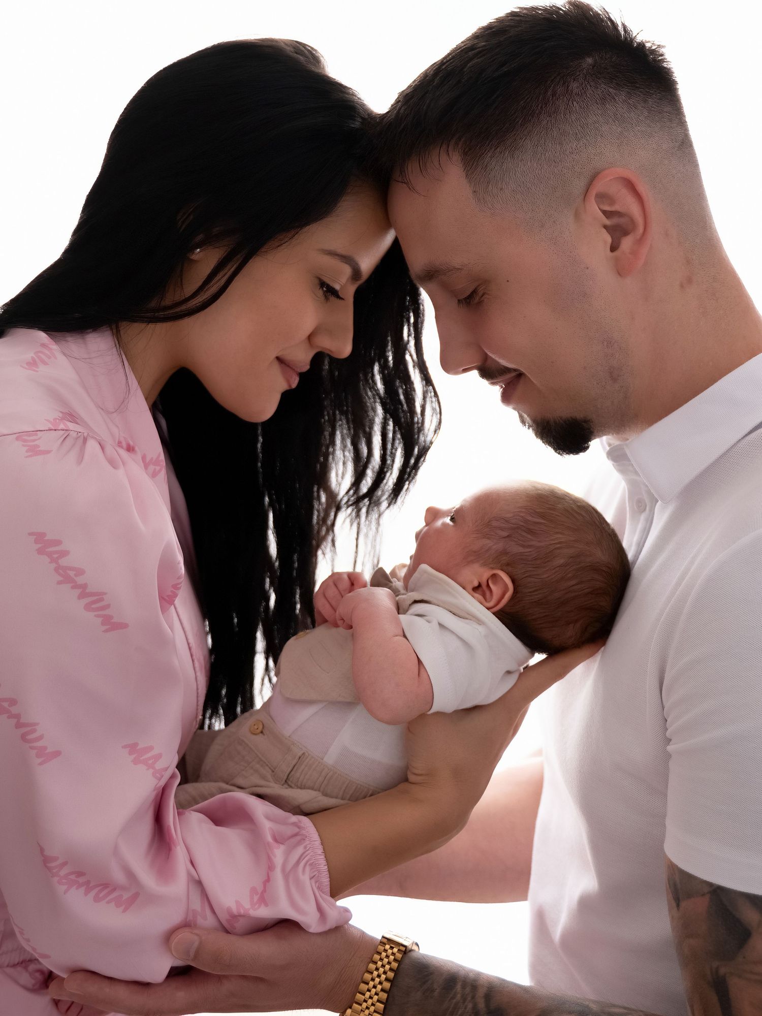 New parents tenderly hold their newborn baby together, mother in pink robe, father with tattoos, sharing an intimate moment.