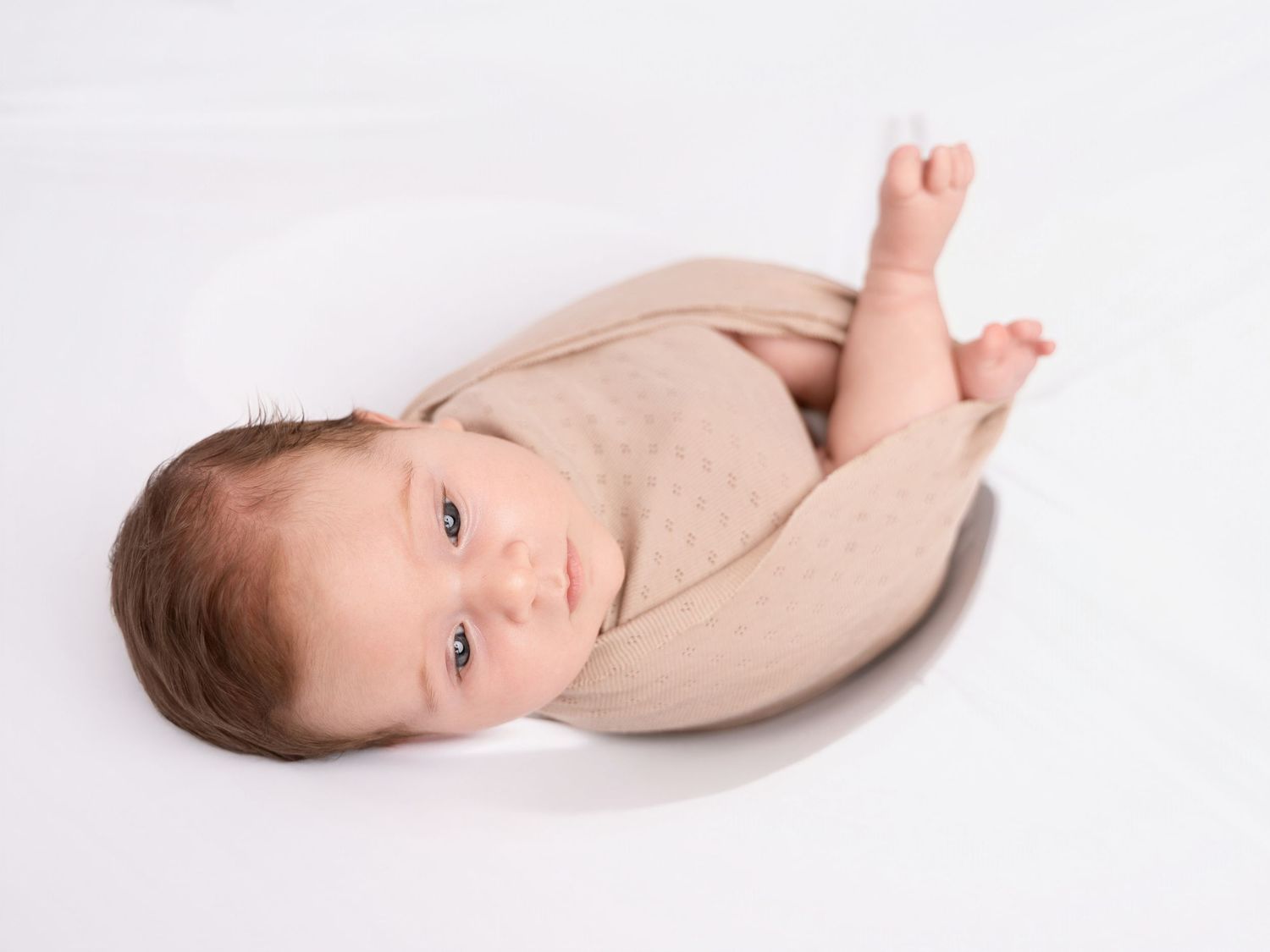 Newborn baby wrapped in beige swaddle lying in white bowl, looking up with bright eyes.