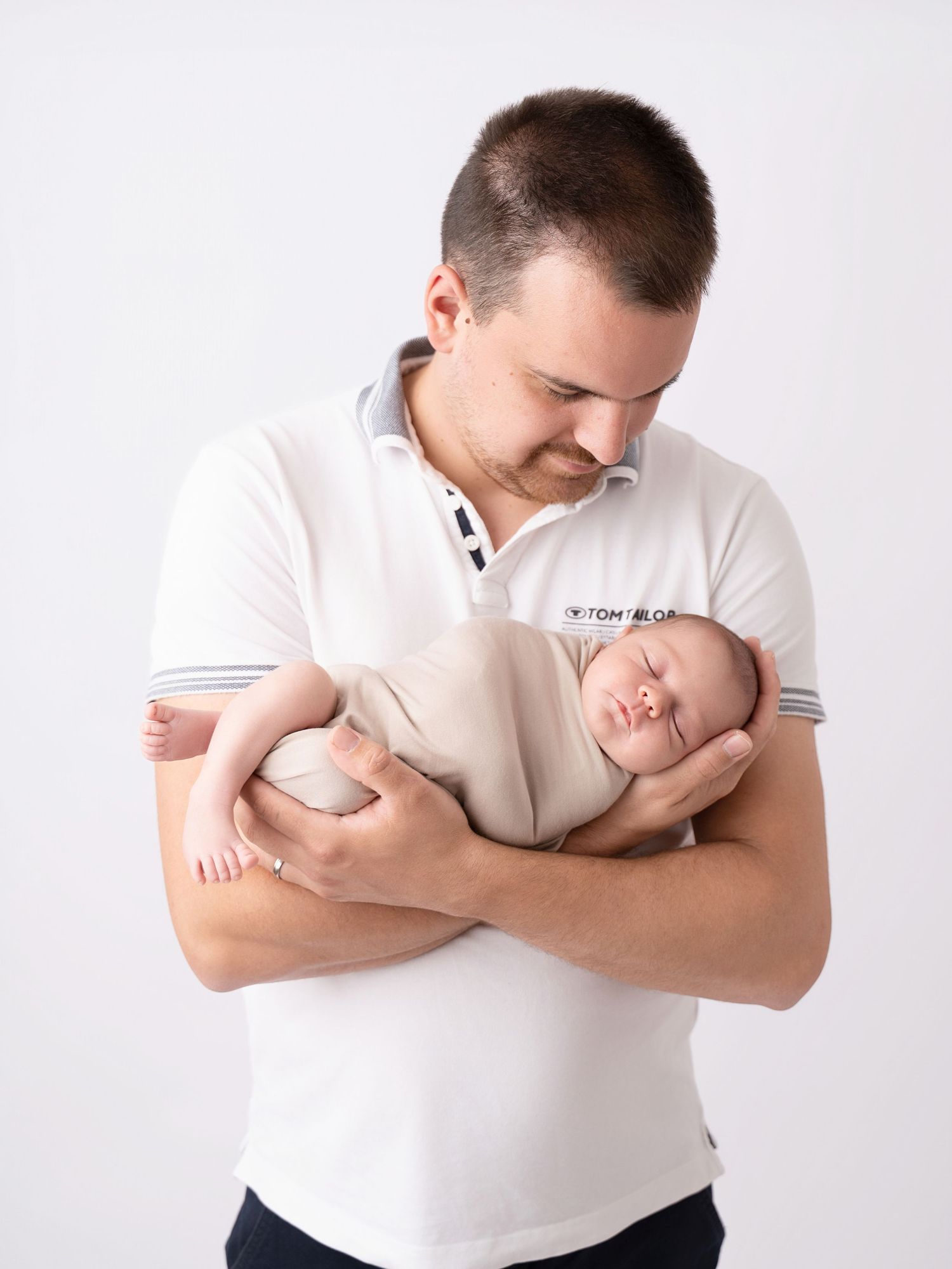 A proud father in a white polo shirt gently cradles his sleeping newborn baby against a white background.