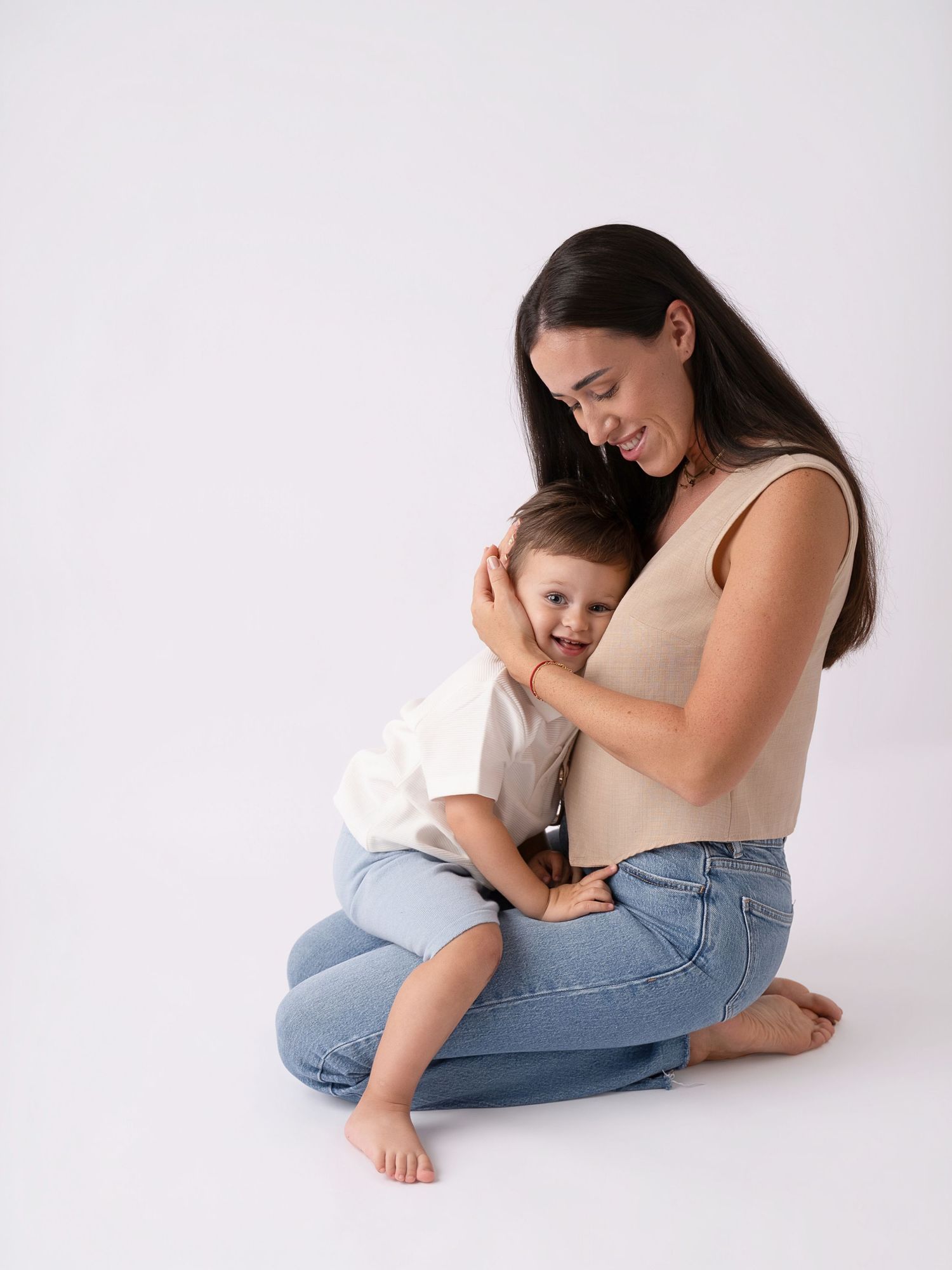 A smiling mother sitting cross-legged on white background, lovingly embracing her happy toddler child.