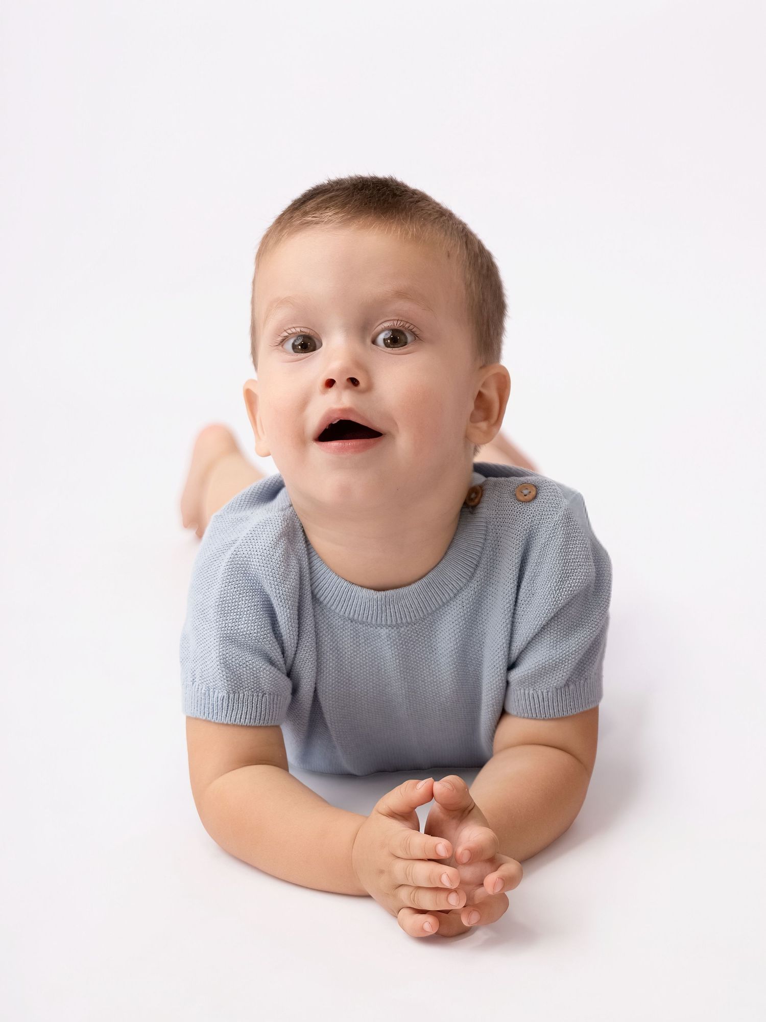 A cute baby boy in a light blue shirt lying on his tummy, looking up with wide eyes and an open mouth.