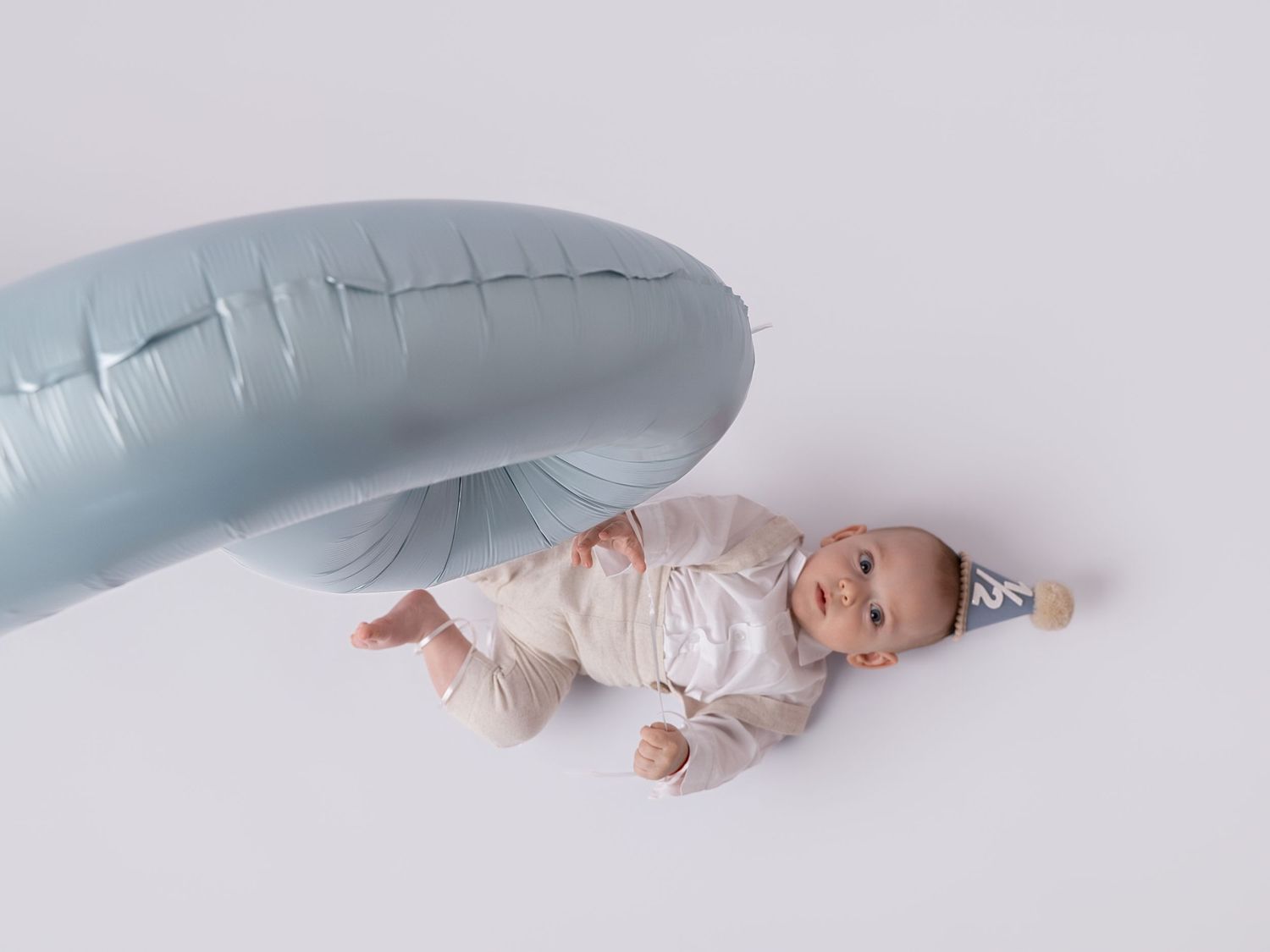 Baby lying next to a light blue inflatable balloon wearing a small party hat on a white background.