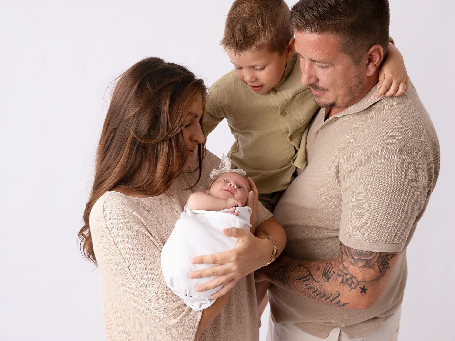 Family of four posing with newborn baby in white studio, parents and toddler admiring the infant.
