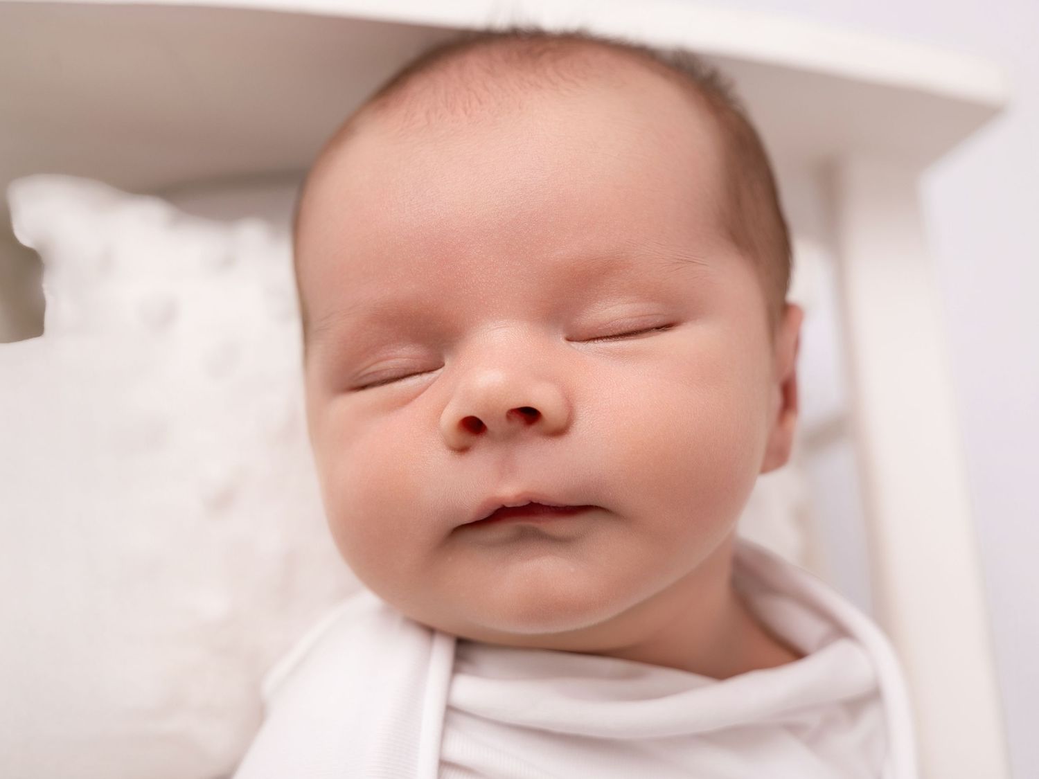 Peaceful newborn baby sleeping soundly in a white crib, swaddled in a soft white blanket.