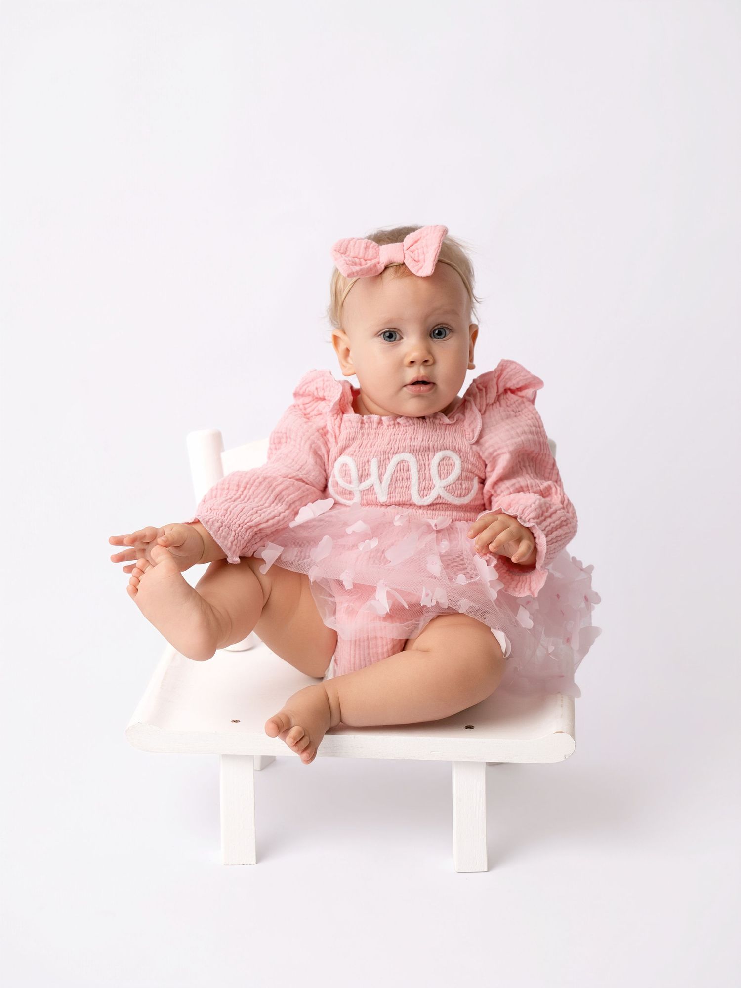 Baby girl in pink 'one' birthday romper with tulle skirt and bow headband sits on white chair.