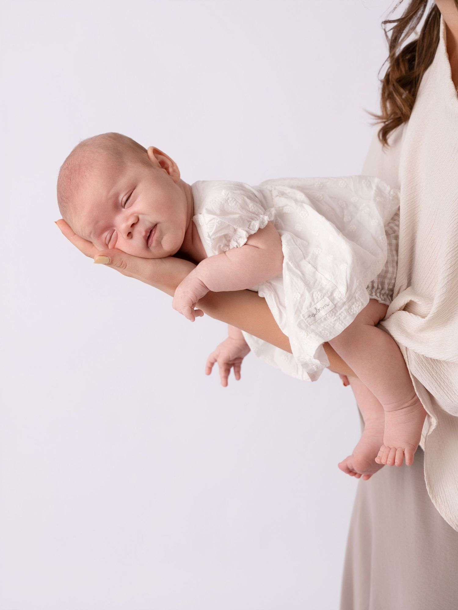 A newborn baby in a white outfit cradled in a mother's hands against a soft white background.