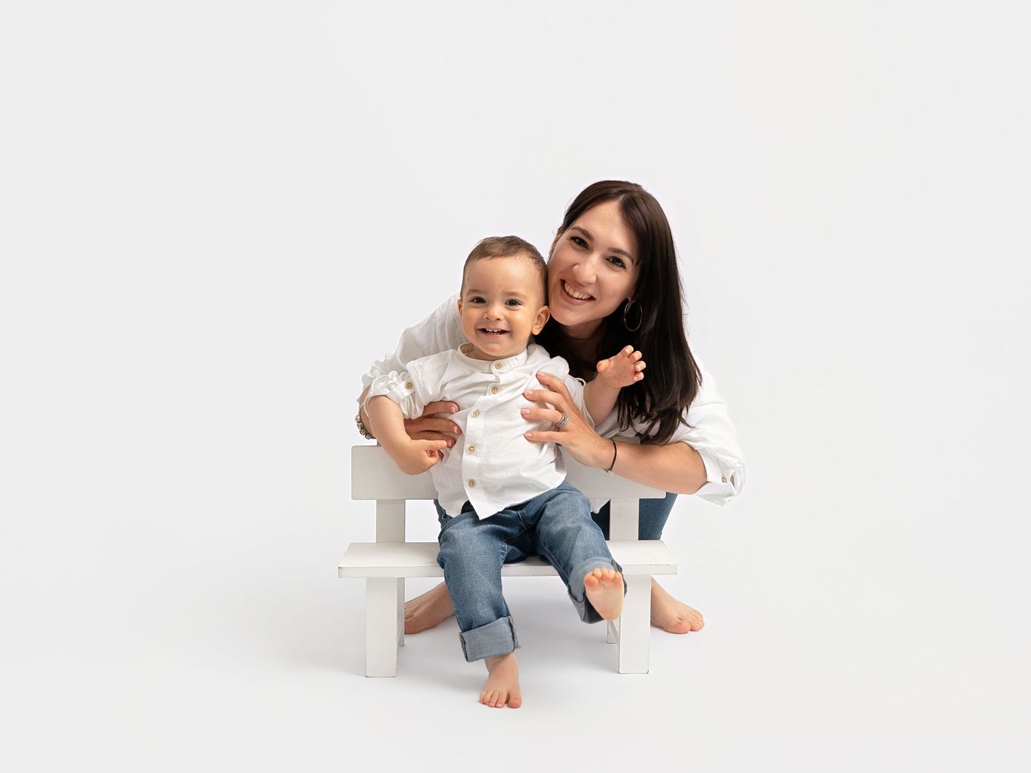 A smiling mother hugs her happy baby boy on a white bench against a clean white background.
