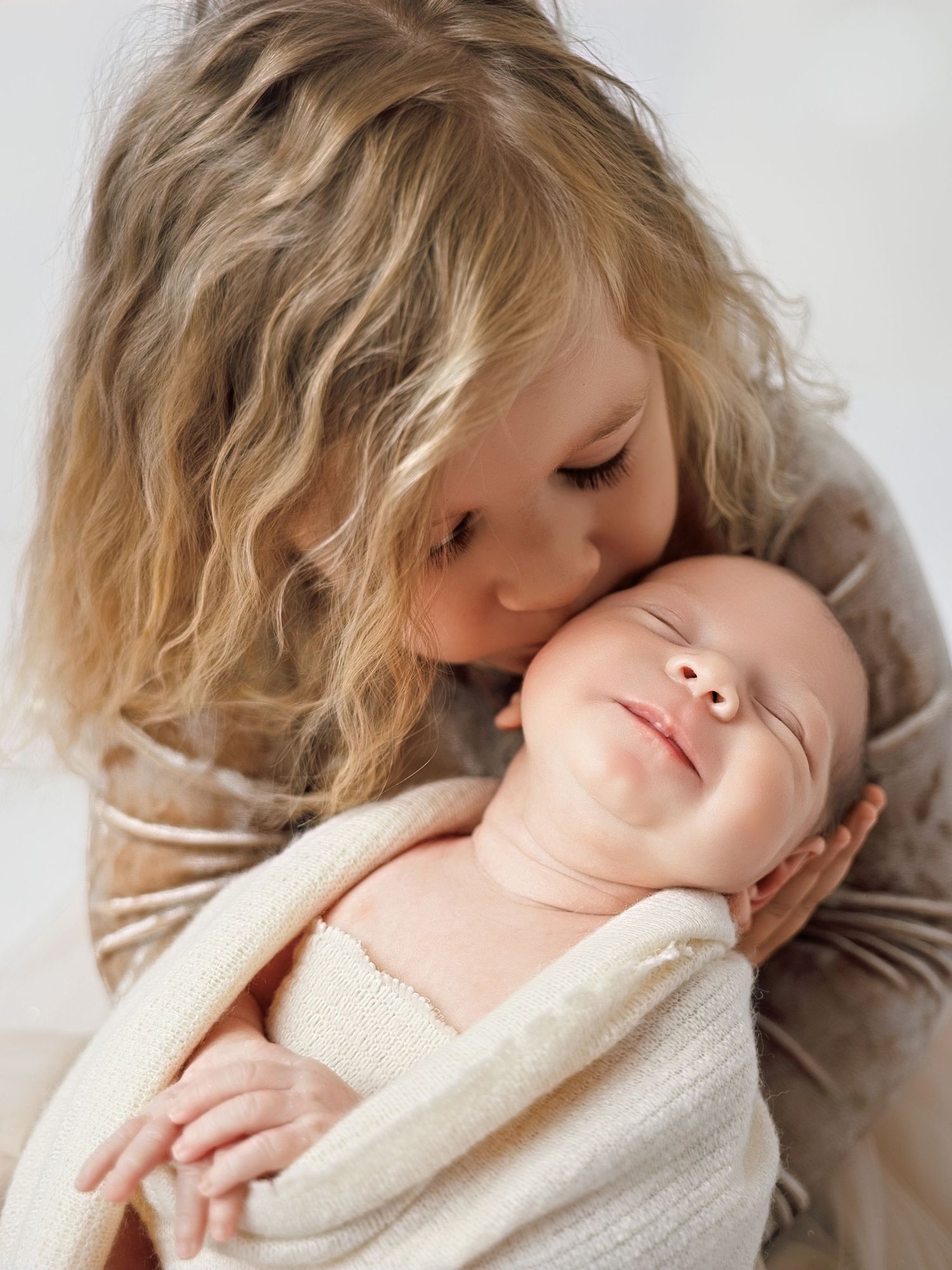 Young girl with curly hair gently kissing a smiling swaddled newborn baby.